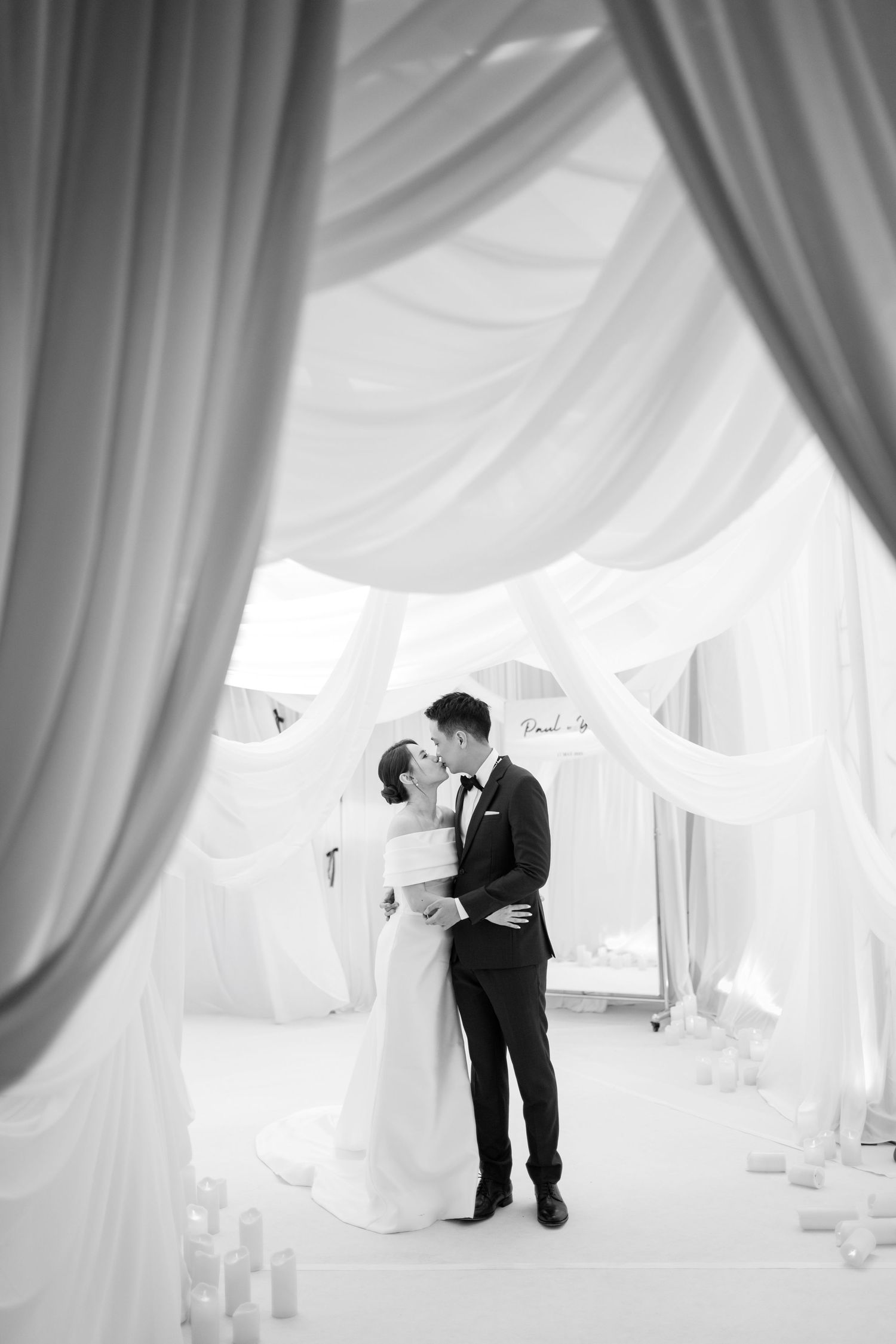 A couple shares a romantic moment in an elegant white draped wedding tent filled with soft light and flowing fabric.