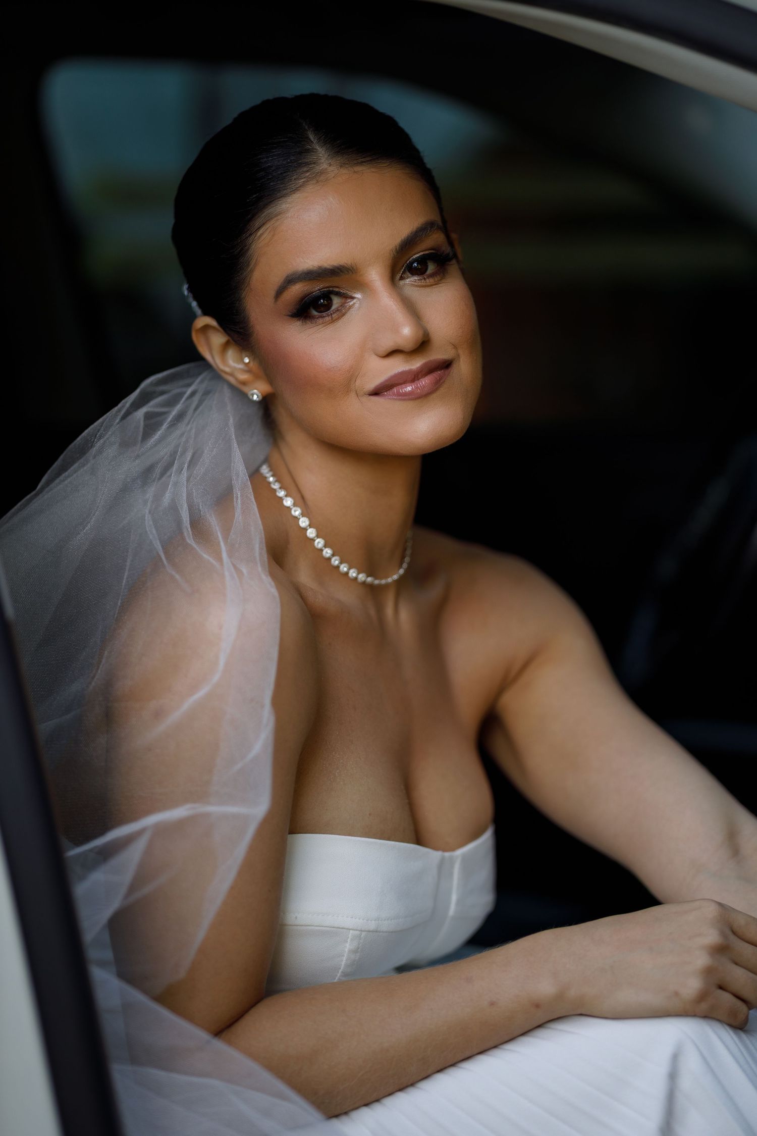 Bride in white wedding dress poses elegantly inside a car with soft lighting.