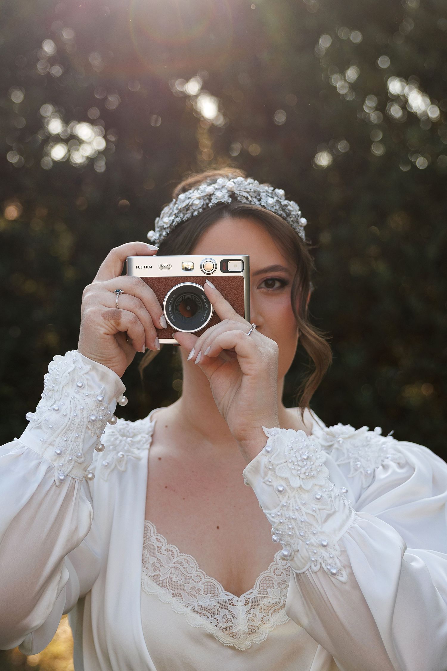 Bride in vintage-style lace dress takes photo with retro camera while wearing crystal headpiece.