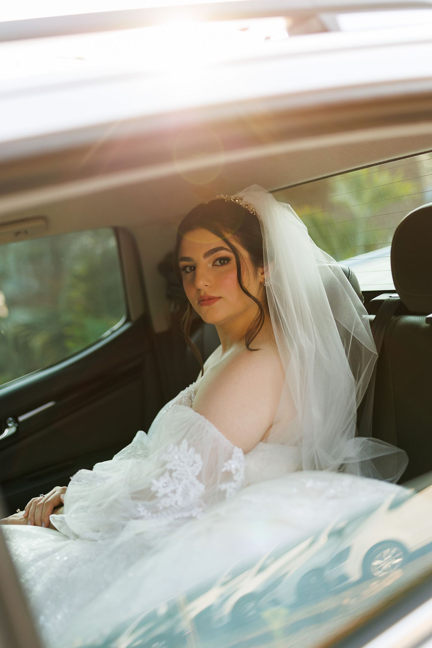A bride in a white lace gown sits in the backseat of a car.