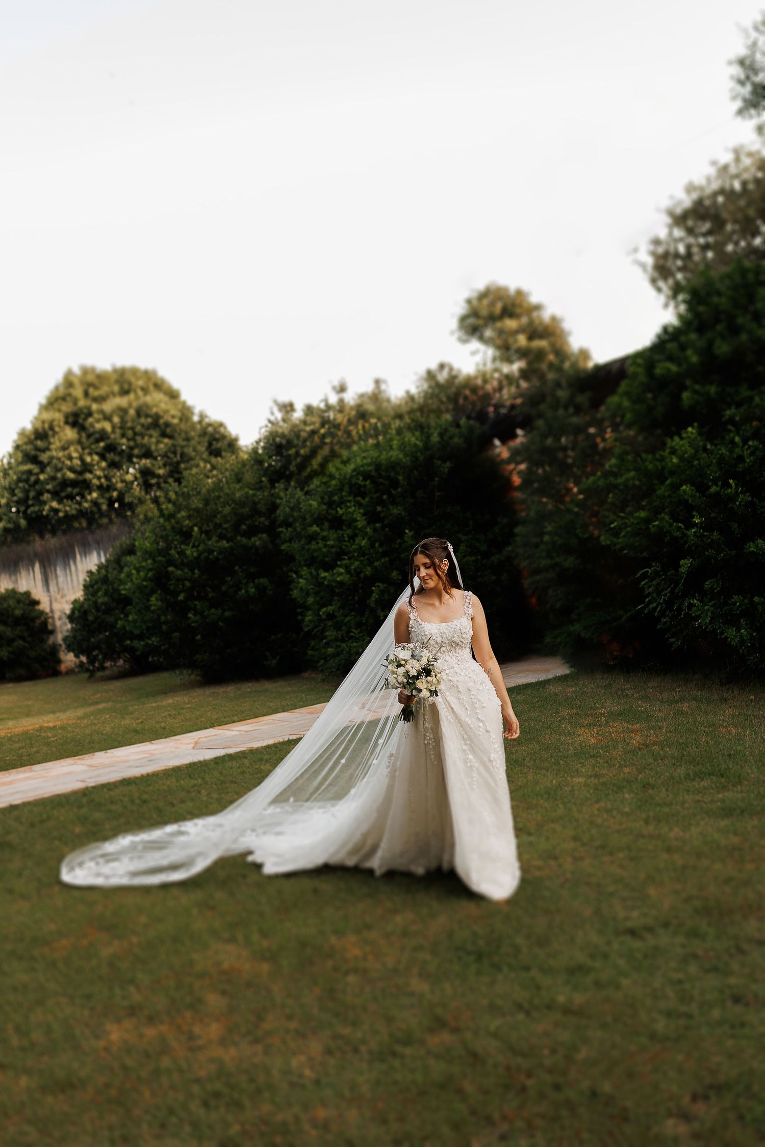 A flowing white wedding dress and veil trail dramatically across a grassy lawn at sunset.