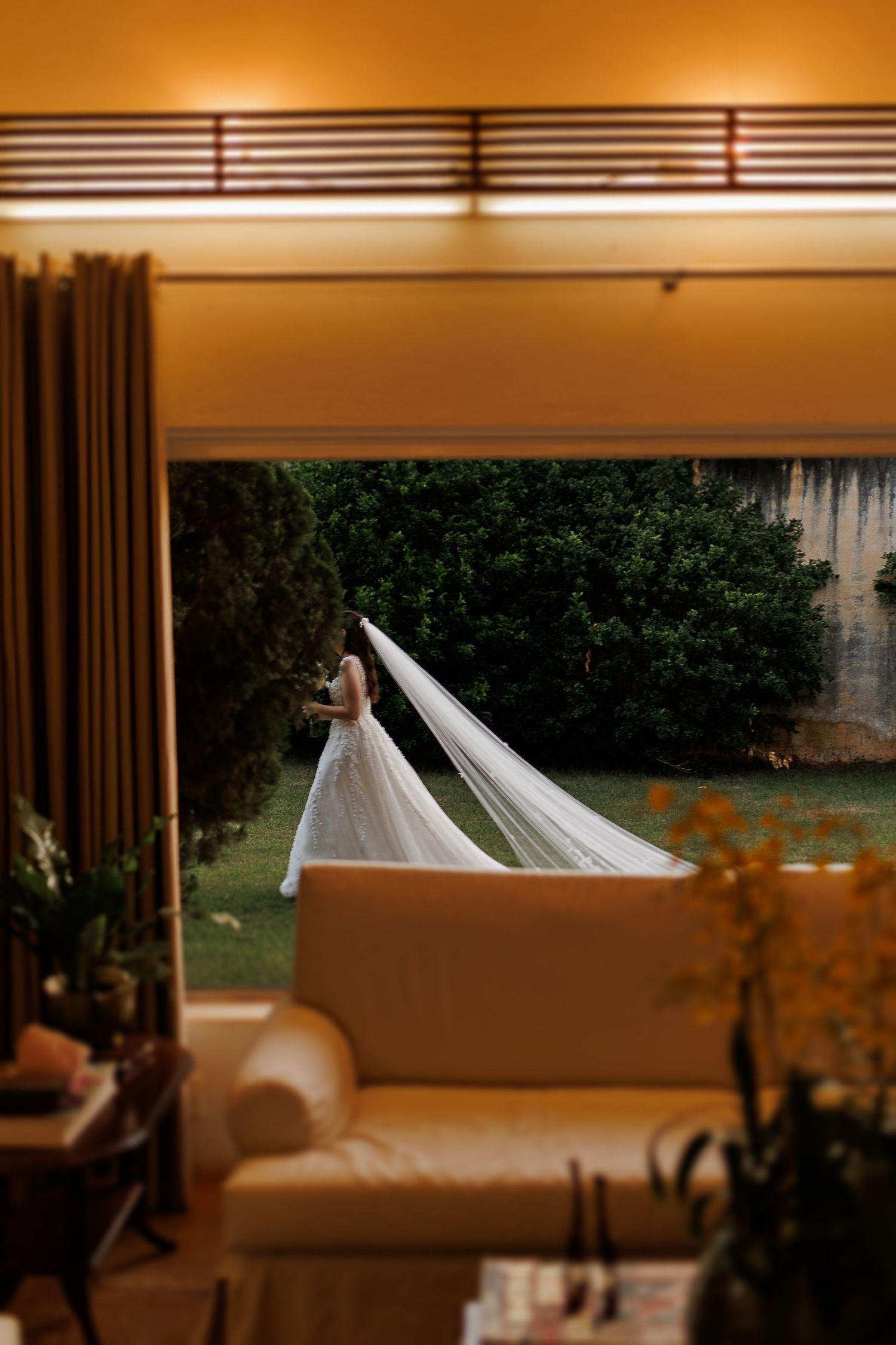 Wedding dress and veil laid out on a couch, bathed in warm ambient lighting through window blinds.