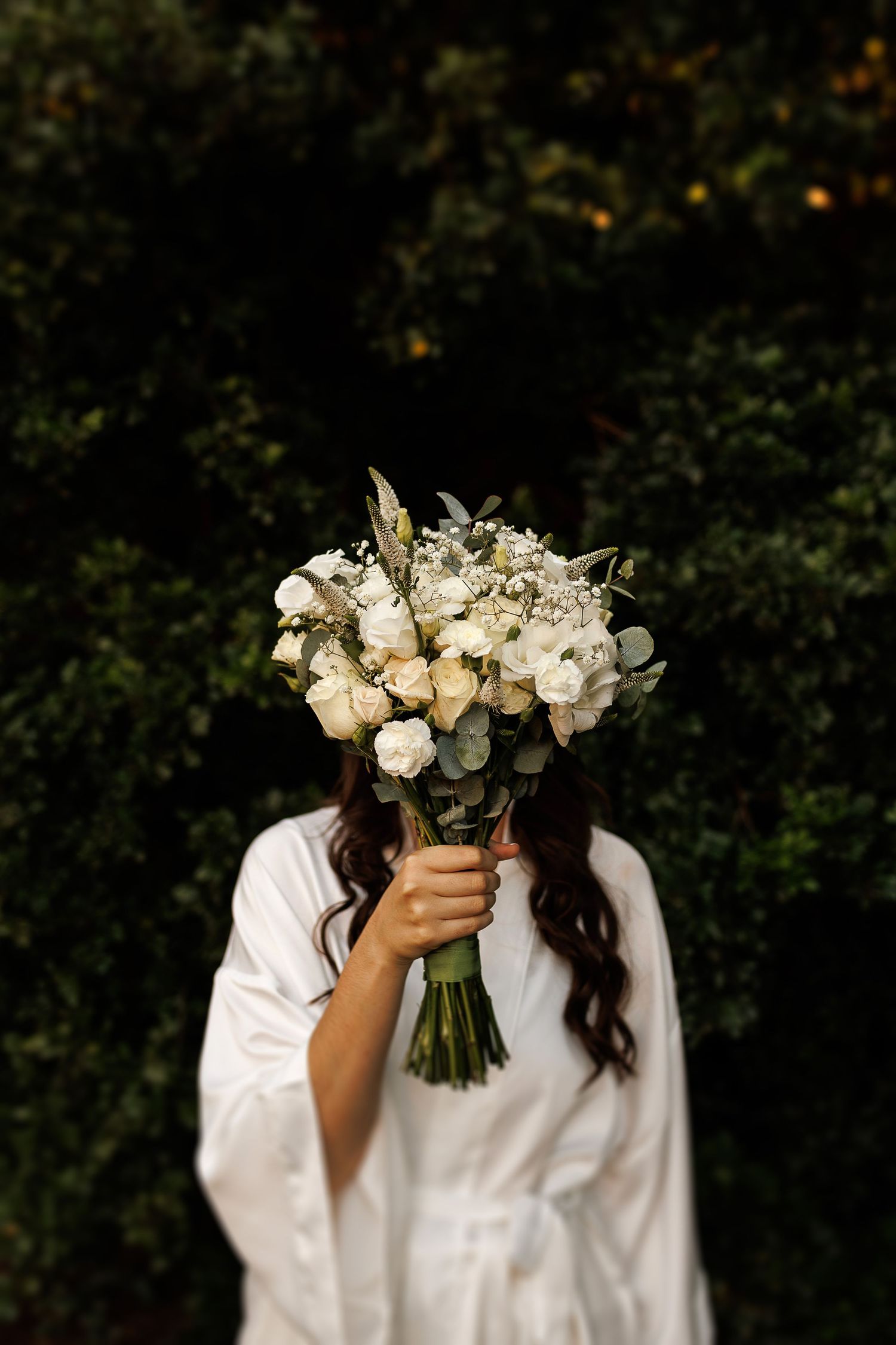 White bridal bouquet with roses and eucalyptus held against dark background.