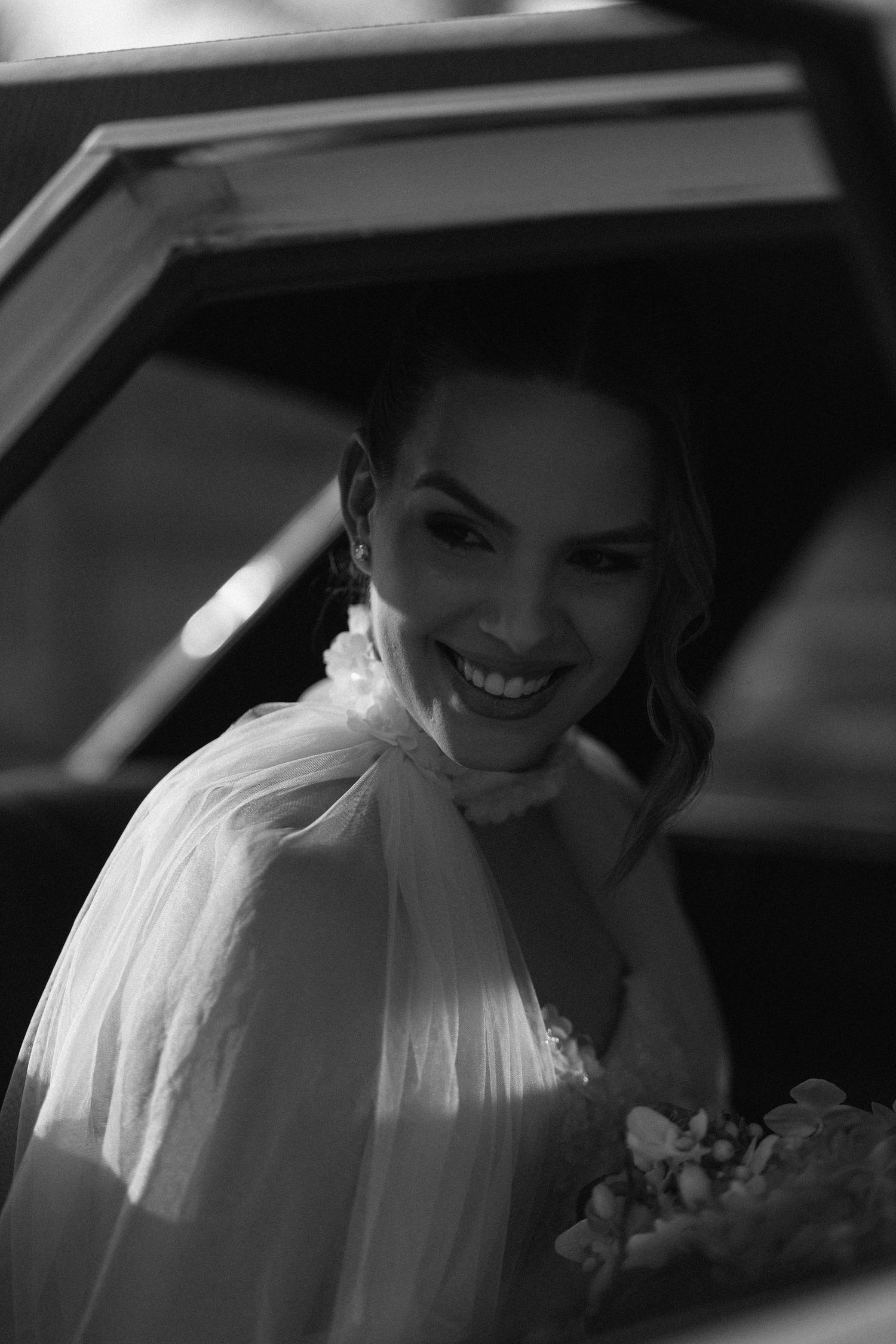 Black and white photo of a bride in a white dress sitting in a car with natural window lighting.