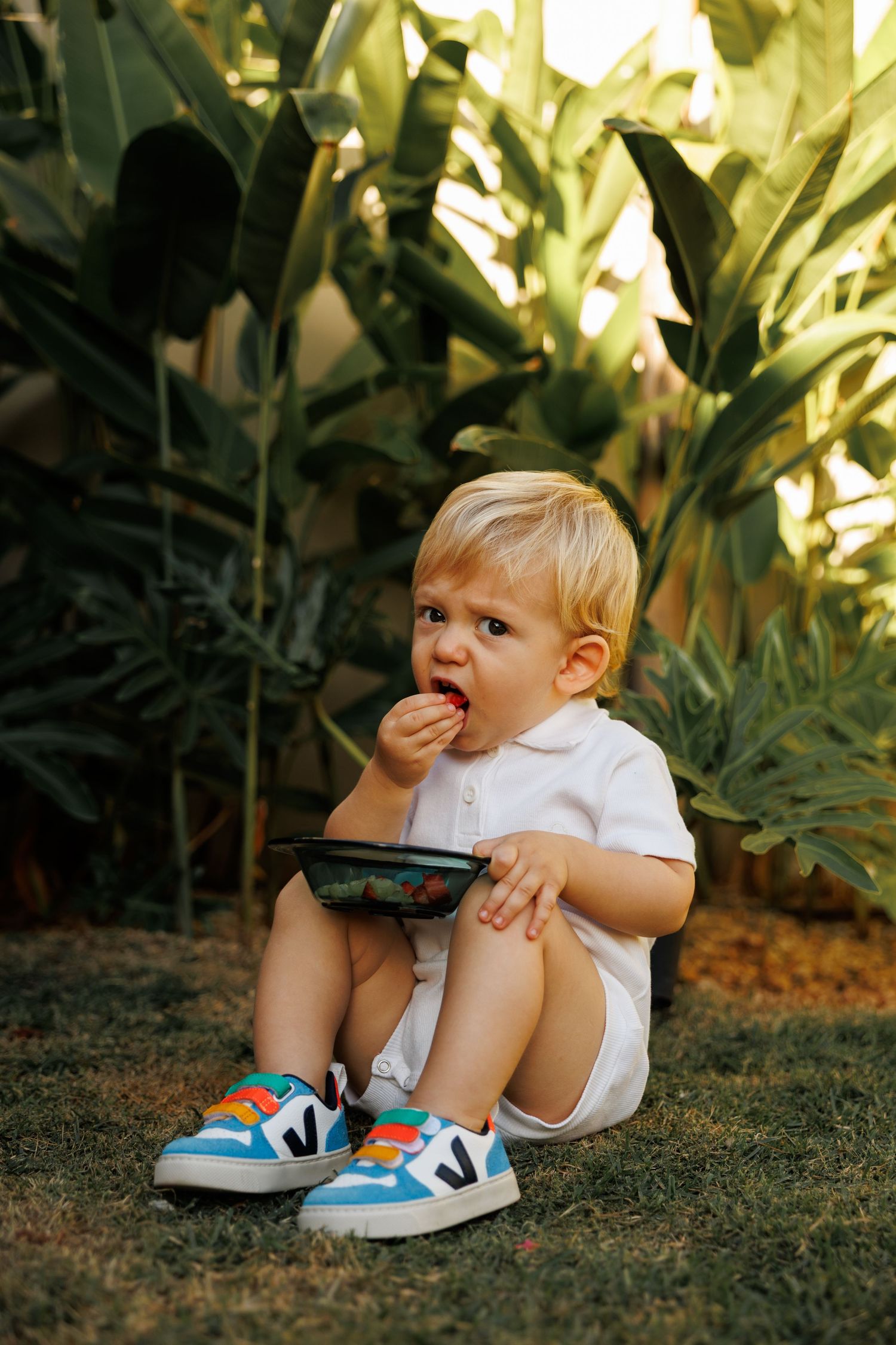 Child in white shirt and blue sneakers sits on grass near tropical plants.