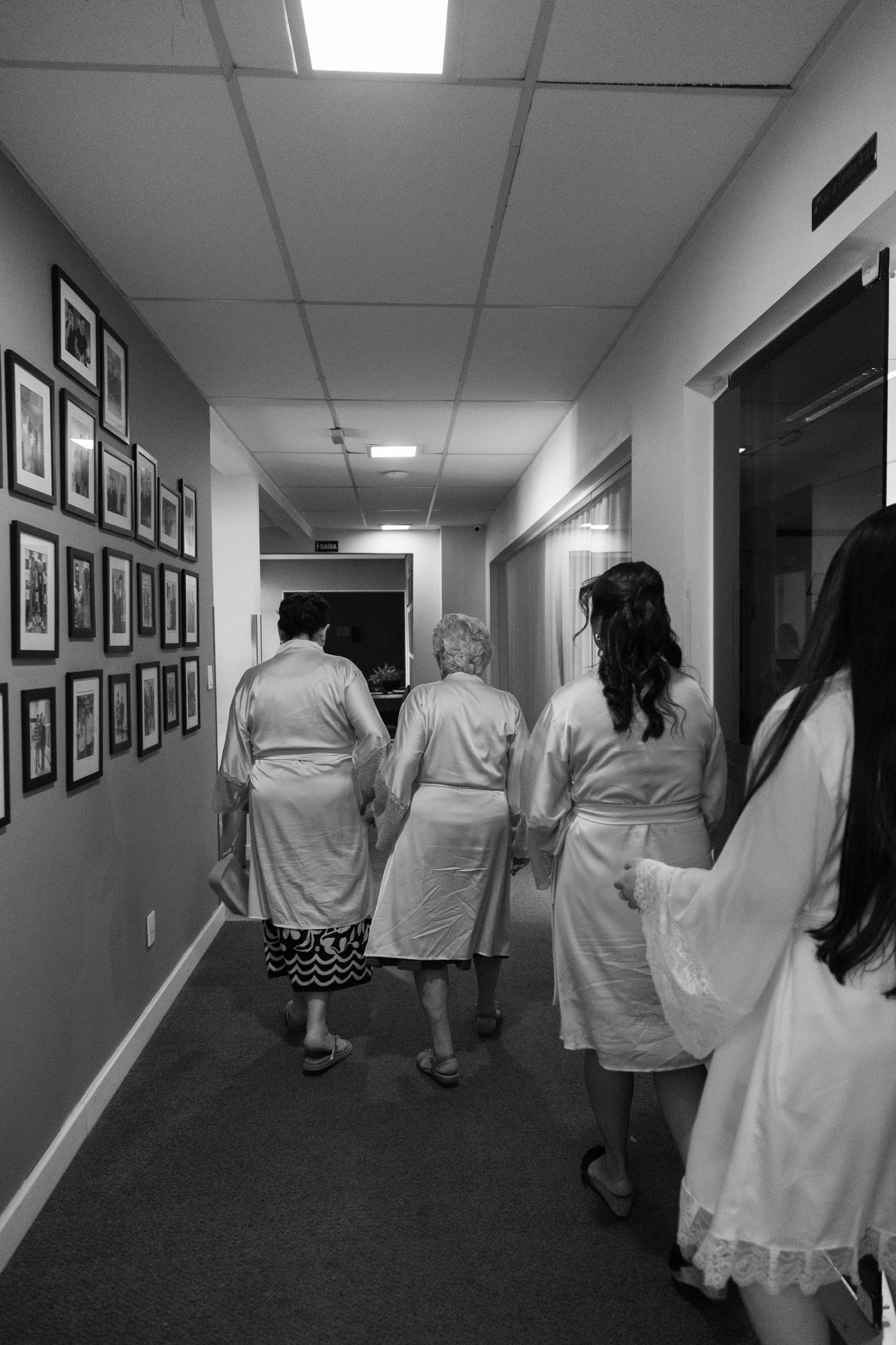A group in white dresses walks down a hallway lined with framed photographs.