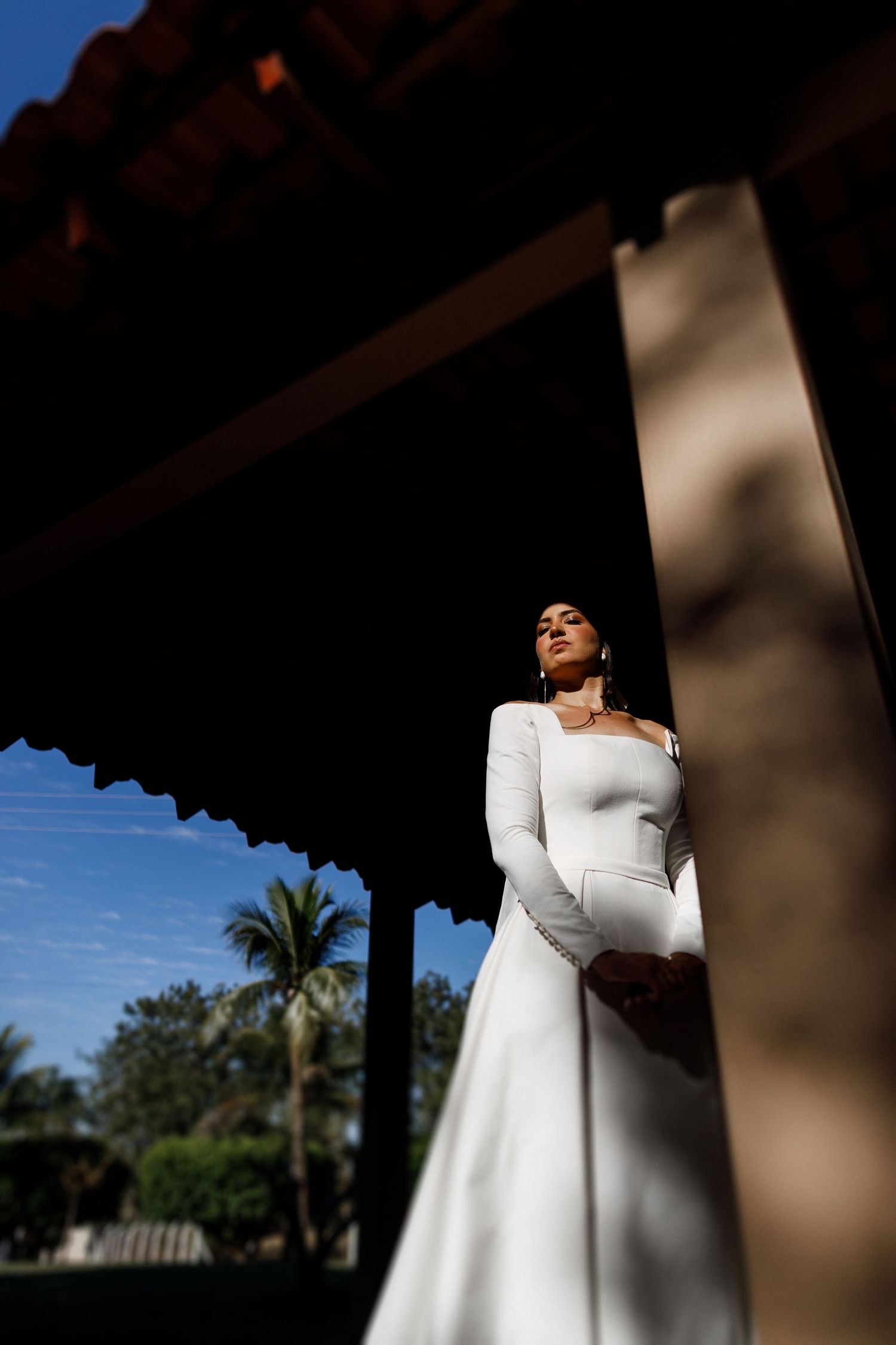 Bride in long-sleeved white dress posing in dramatic lighting with palm trees in background.