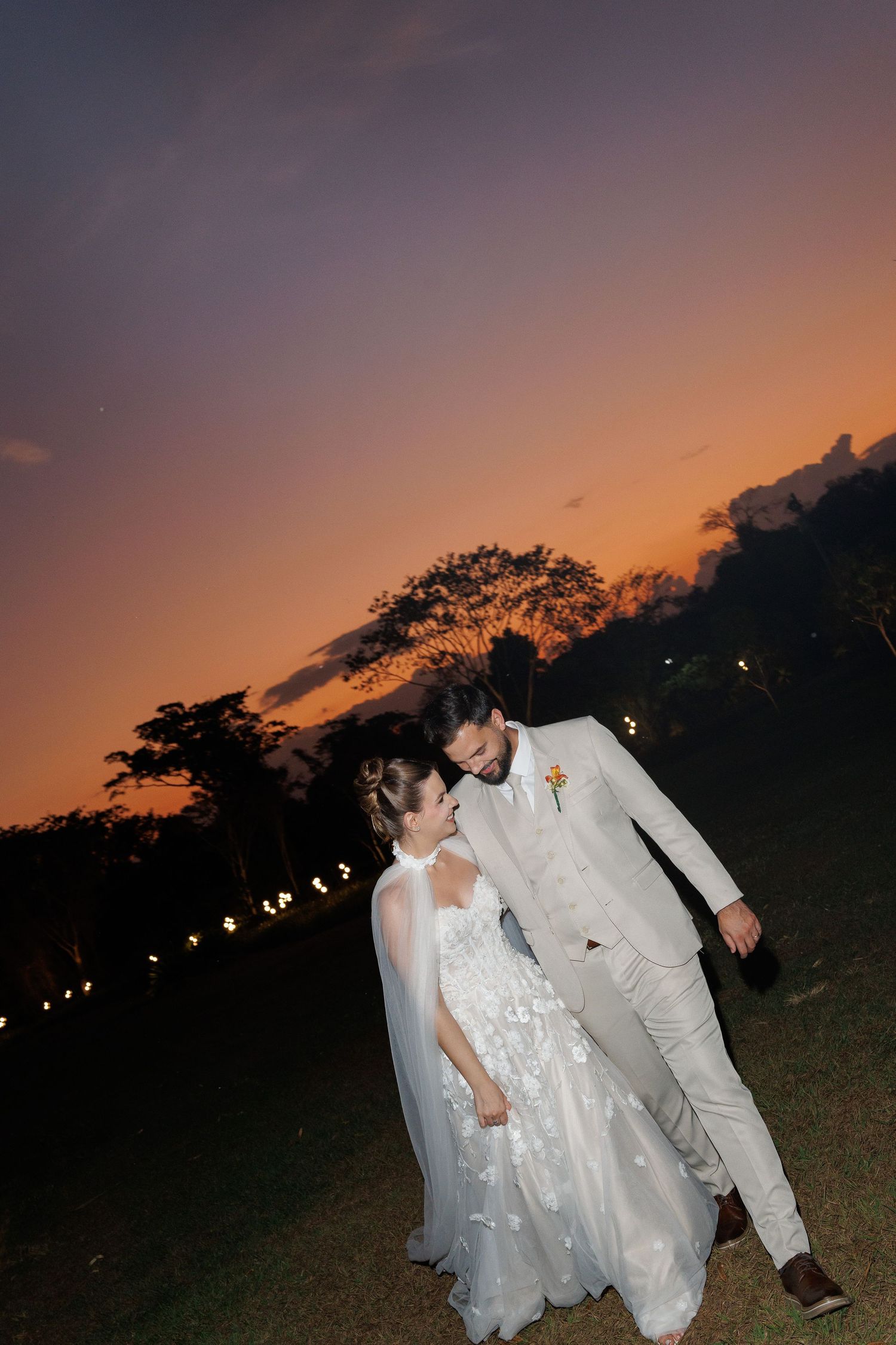 Newlyweds in white attire dance together at dusk with string lights in the background.
