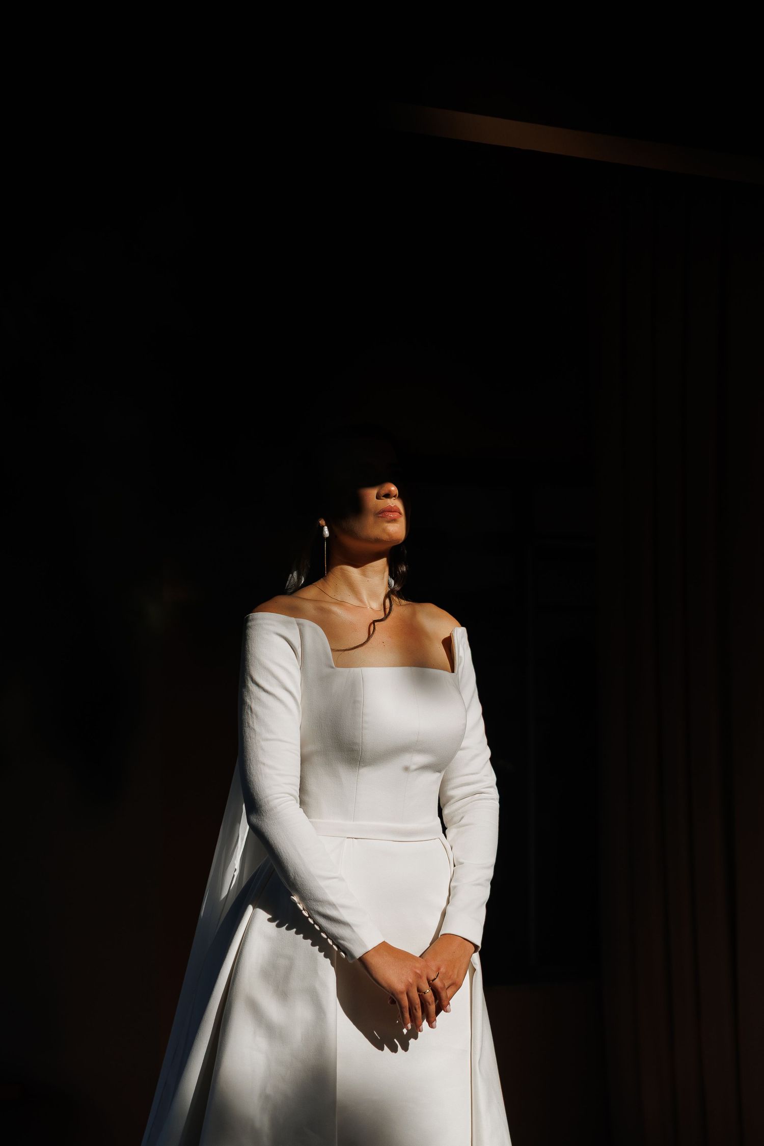 Elegant long-sleeved white wedding dress captured in dramatic window light.