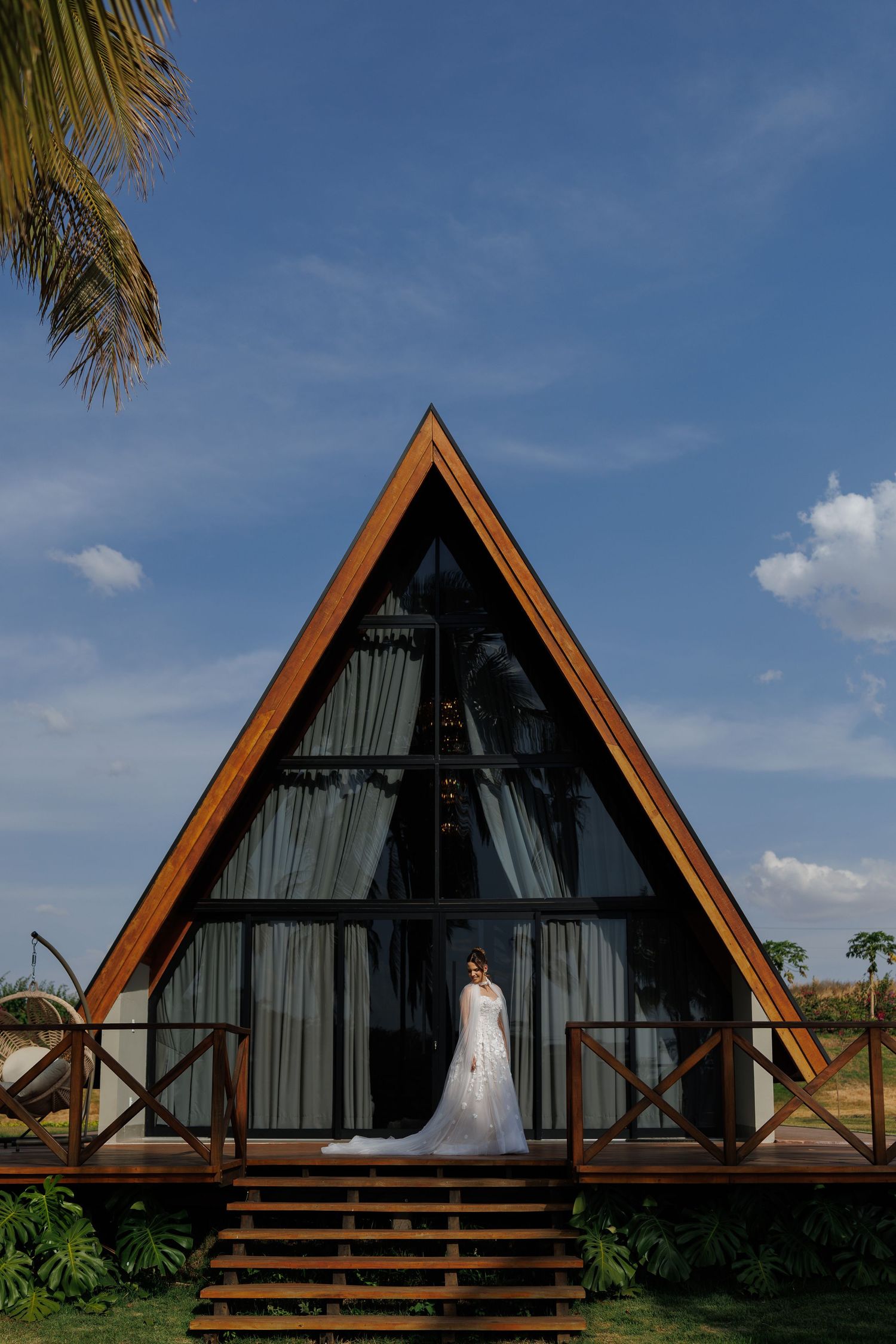 A-frame wooden cabins with dark exteriors stand against a blue sky with palm trees in a tropical setting.