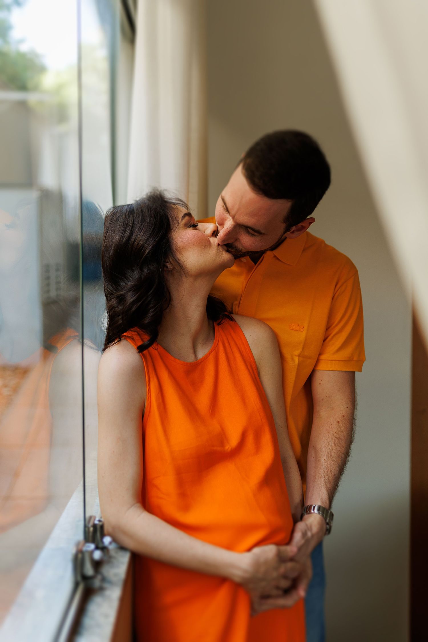 Couple in orange and dark clothing sharing an intimate moment by a window.
