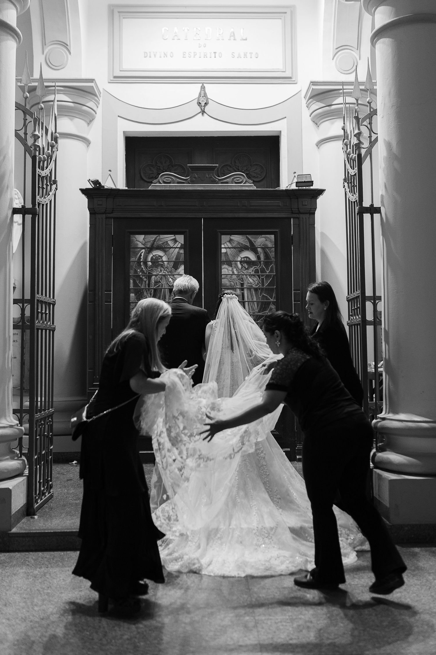 Silhouettes help arrange a bride's wedding dress train in an elegant indoor hallway.