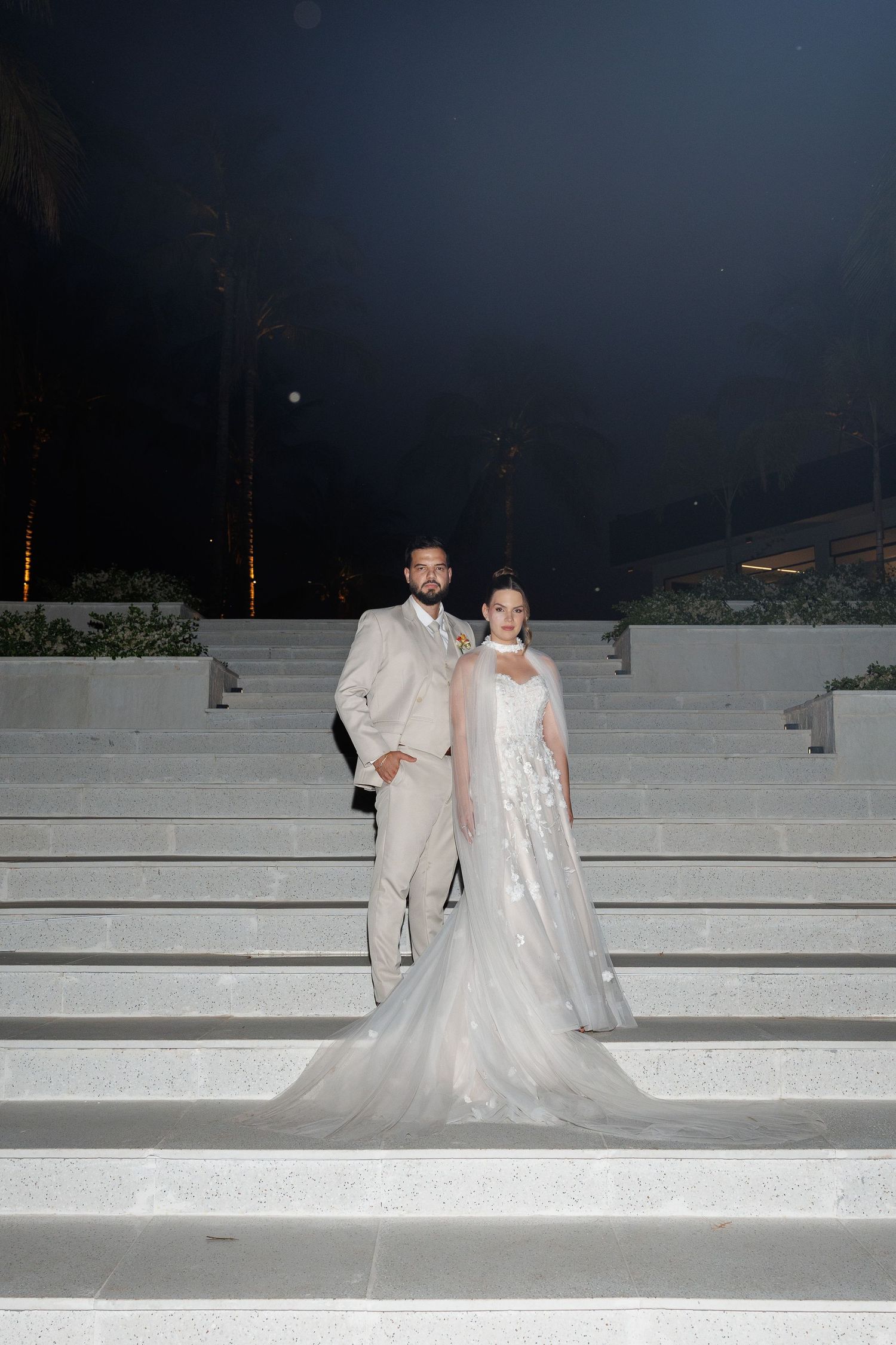 Wedding couple poses on white concrete steps at night in formal attire.