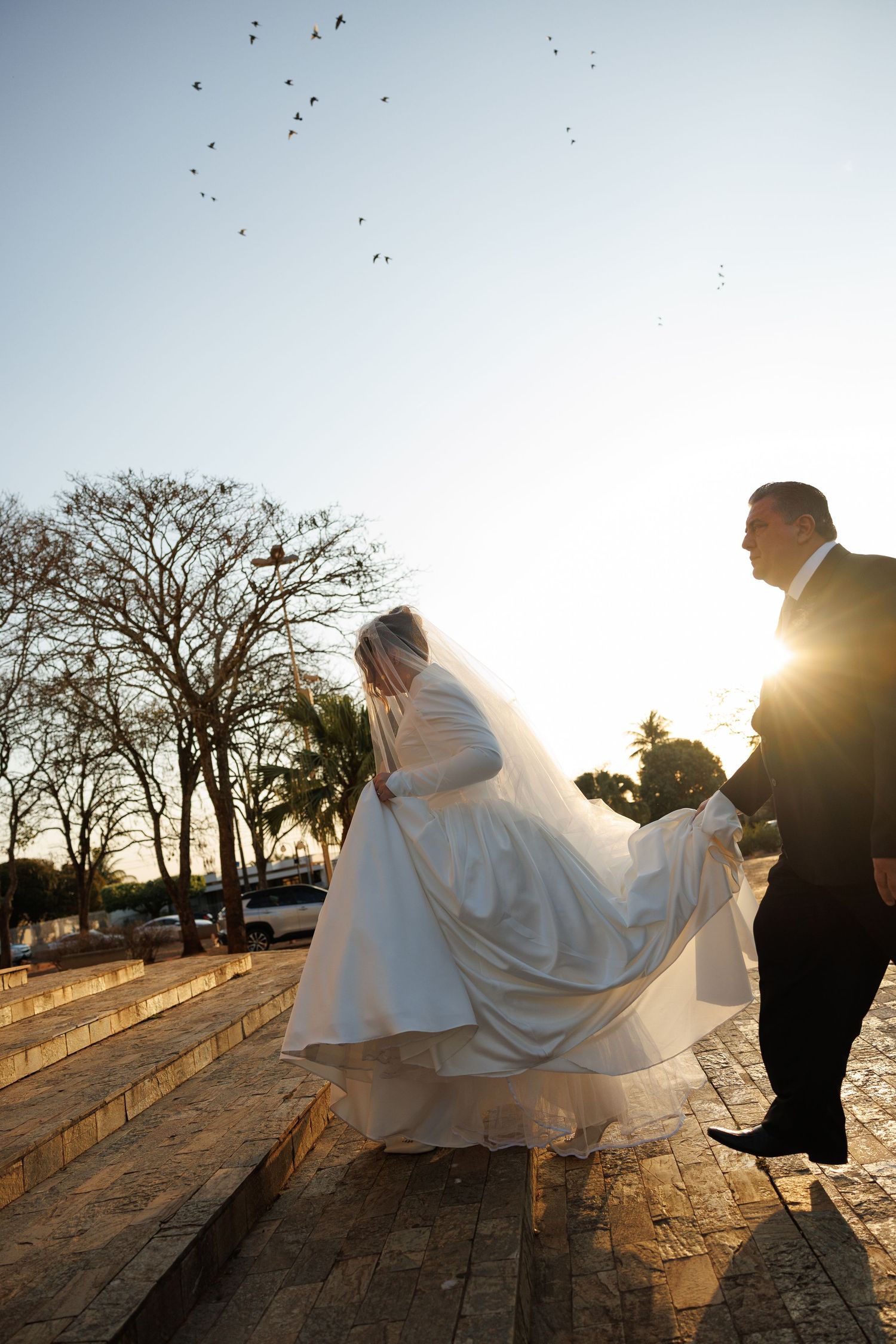 Silhouetted couple in wedding attire walking along a path at sunset with birds flying overhead.