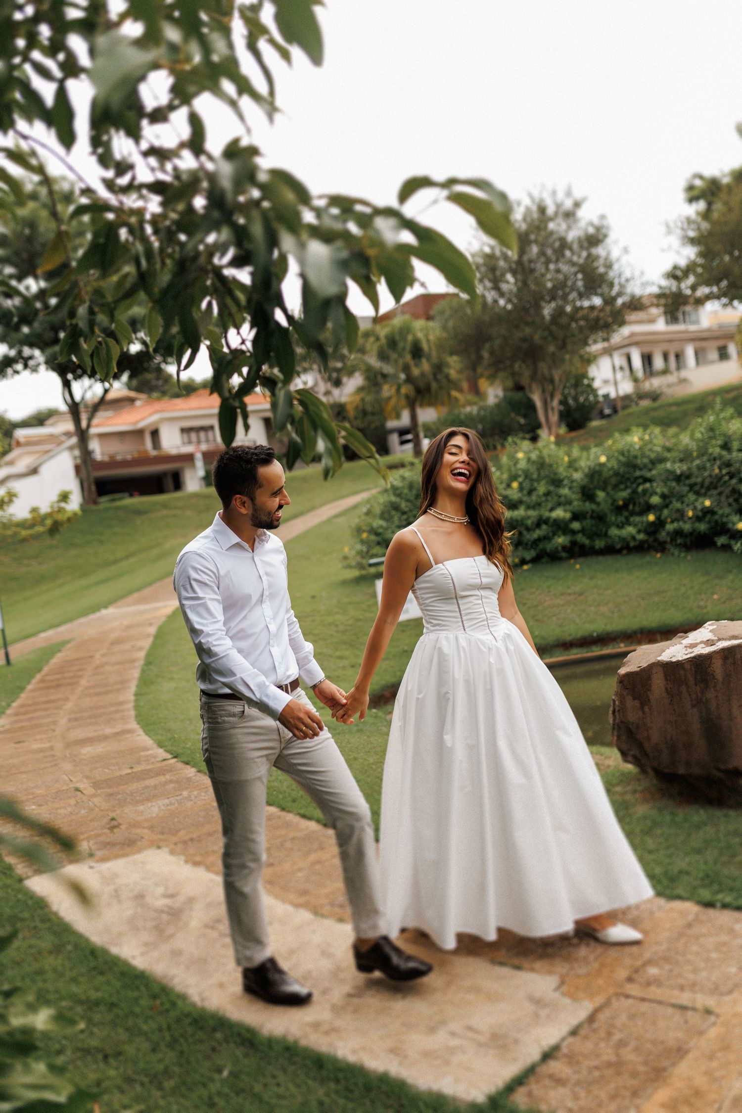 Couple in wedding attire walking and laughing along a curved garden path.