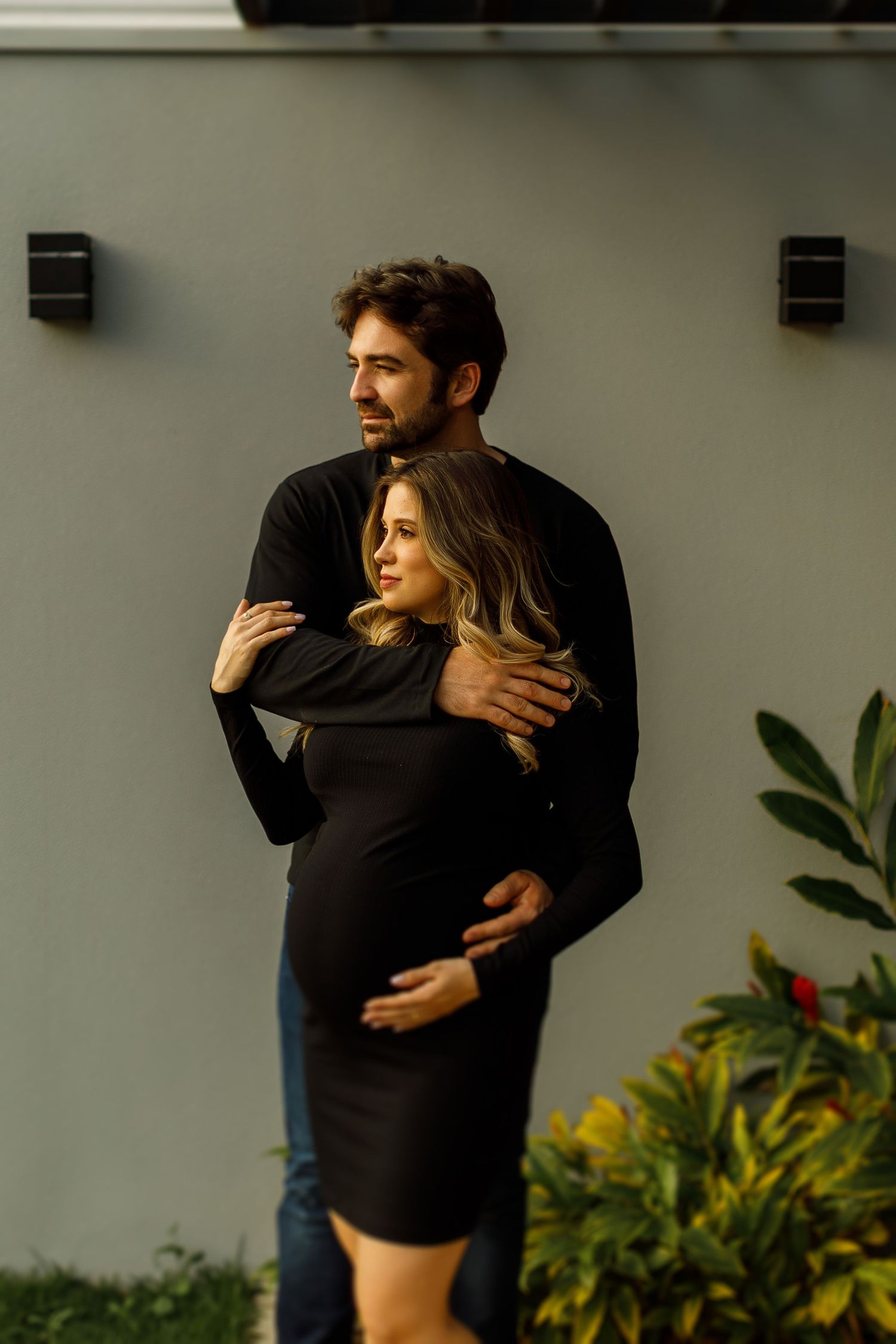 Couple embracing outdoors with greenery and modern architecture in background.