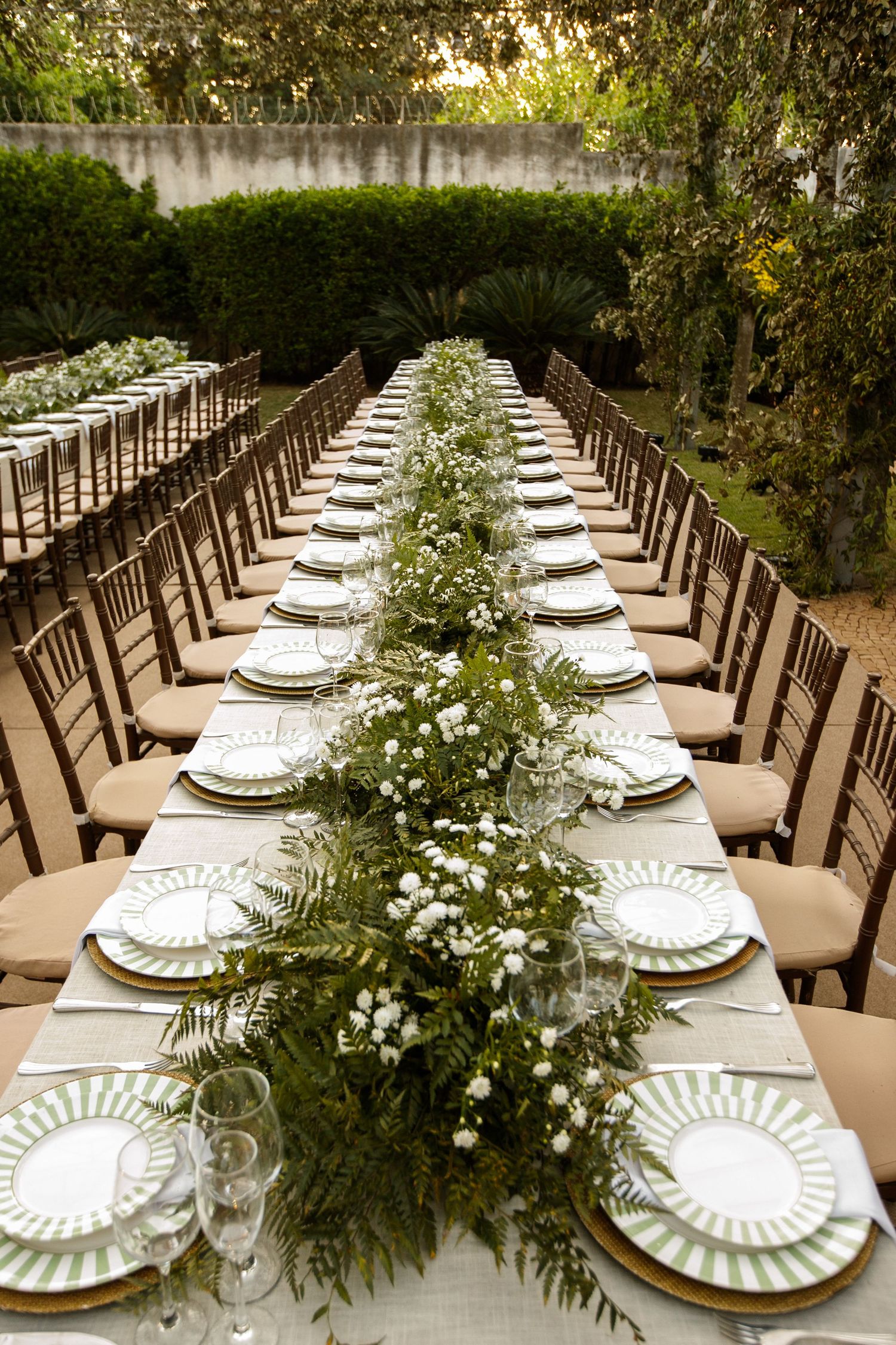 Long wedding reception table set with white plates and lush greenery centerpieces under natural lighting.