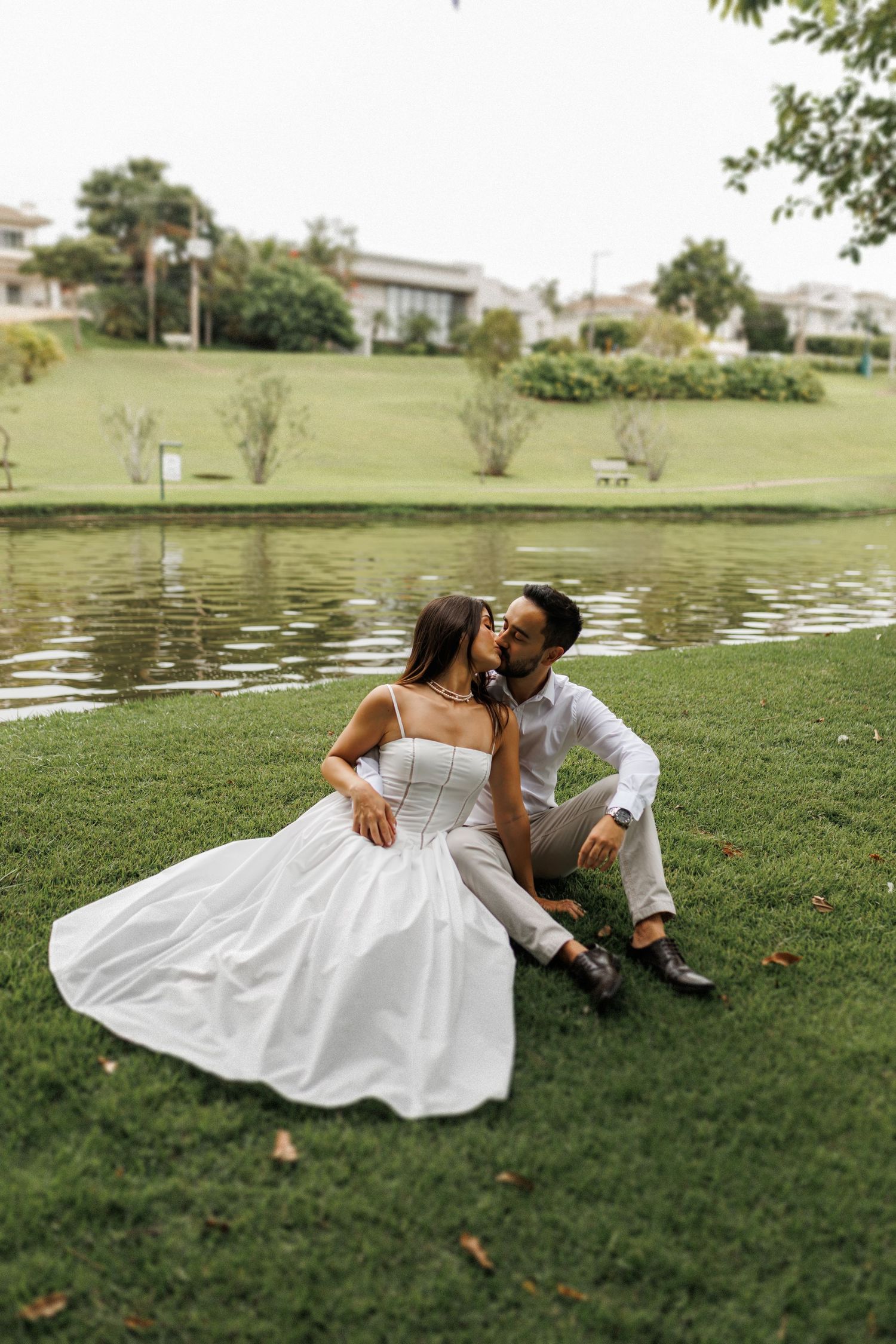 Romantic couple in white outfits lounging on grass near a pond.