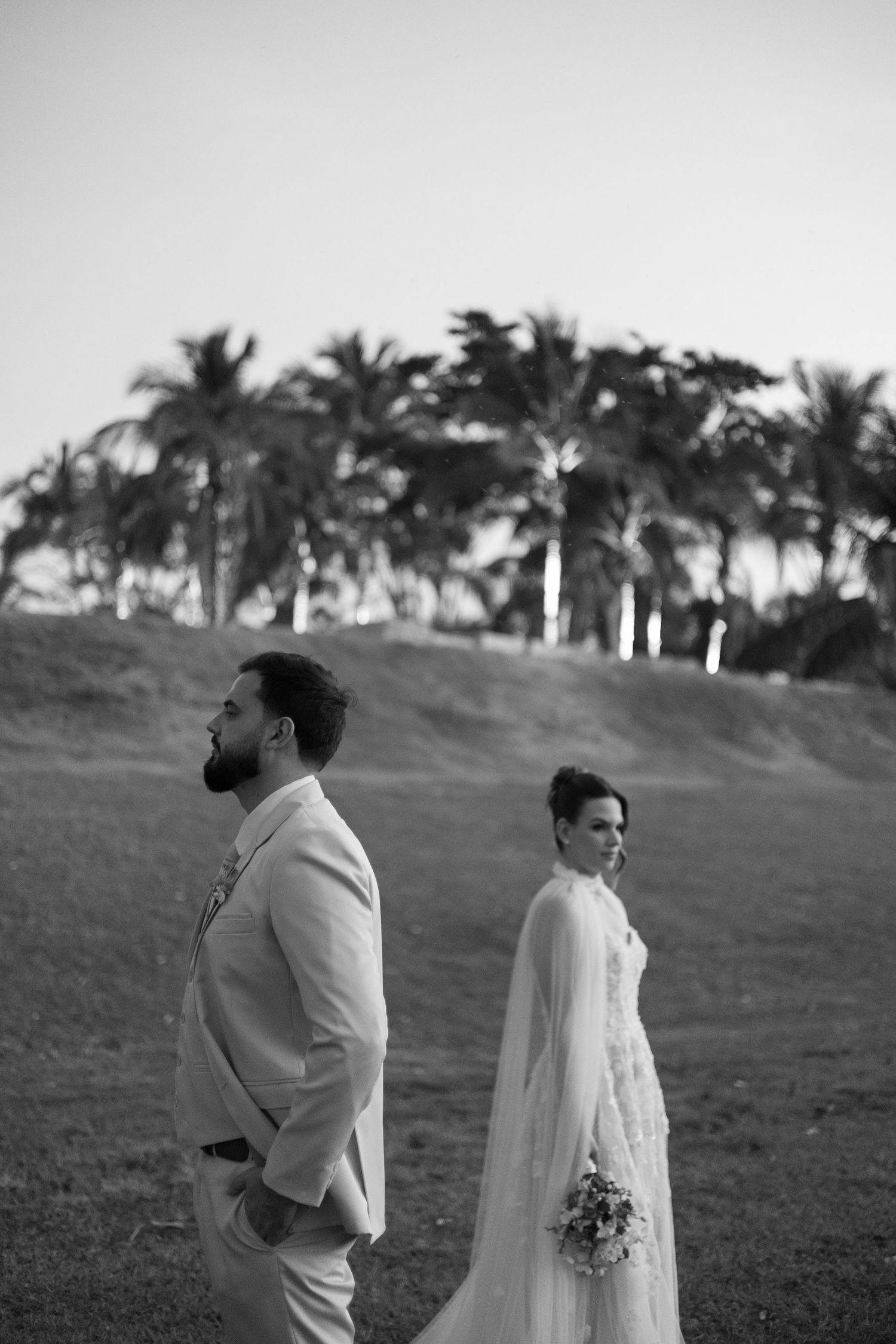 A black and white portrait of newlyweds standing in a palm tree-lined field.