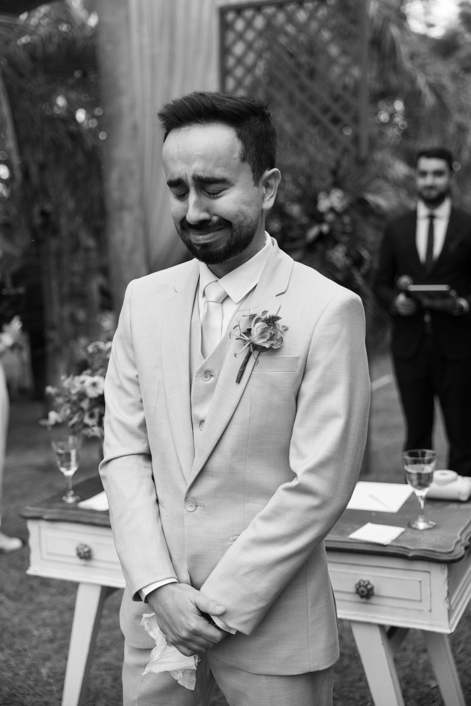 A groom in a light colored suit smiles during an outdoor wedding ceremony.