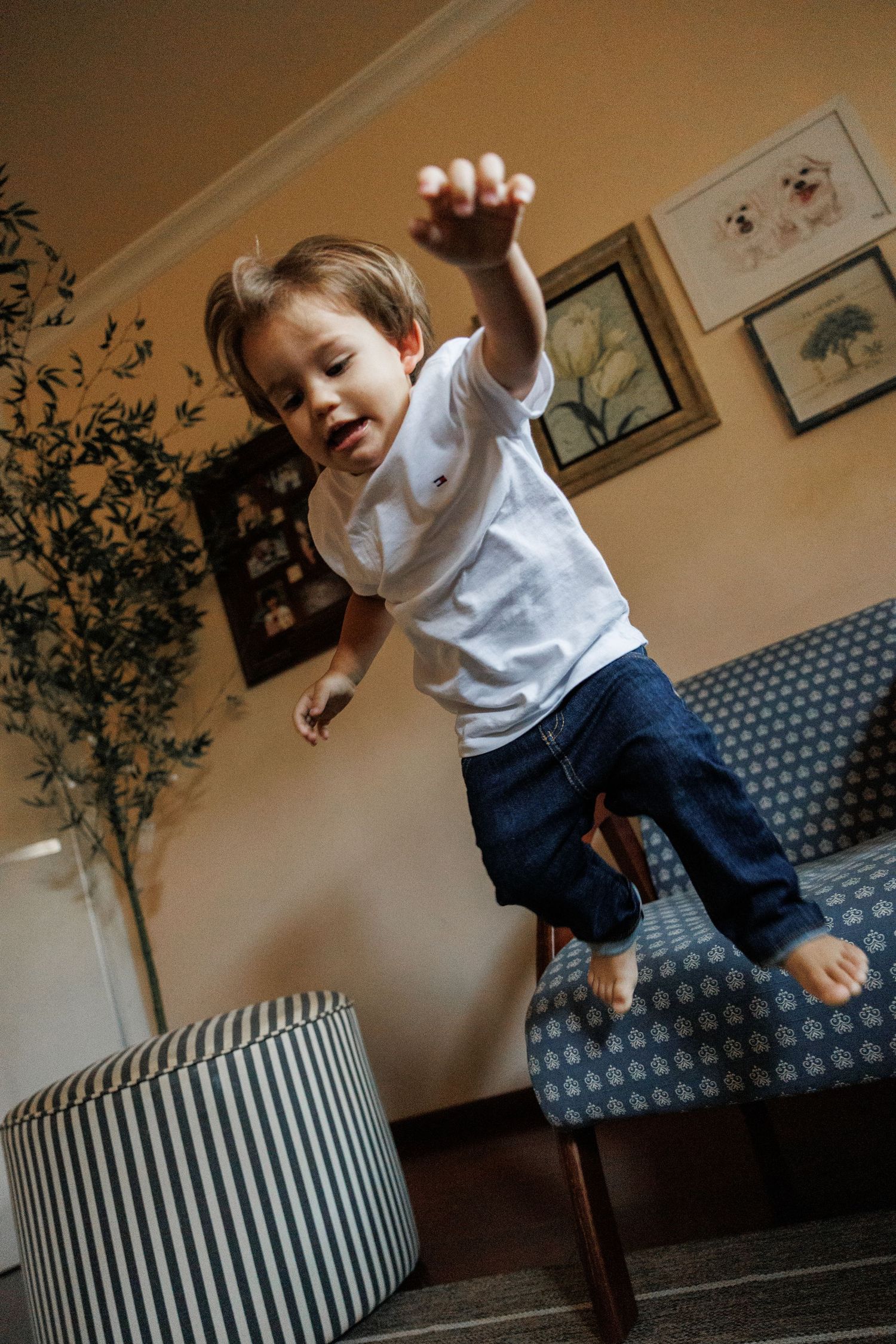 Child in white shirt and jeans jumping playfully on a striped ottoman in a living room setting.