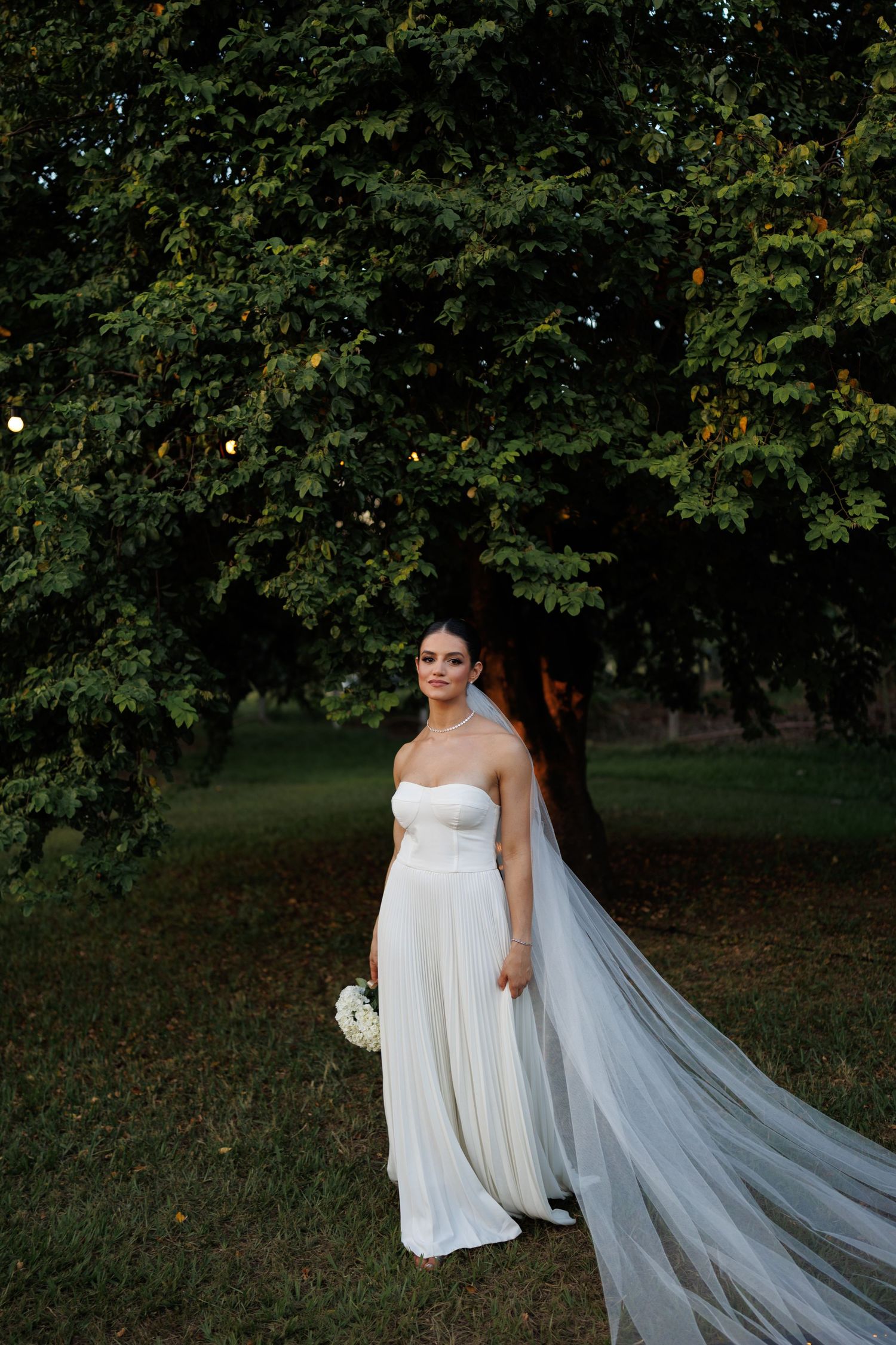 Bride in flowing white gown and veil walks gracefully through a tree-lined garden setting.