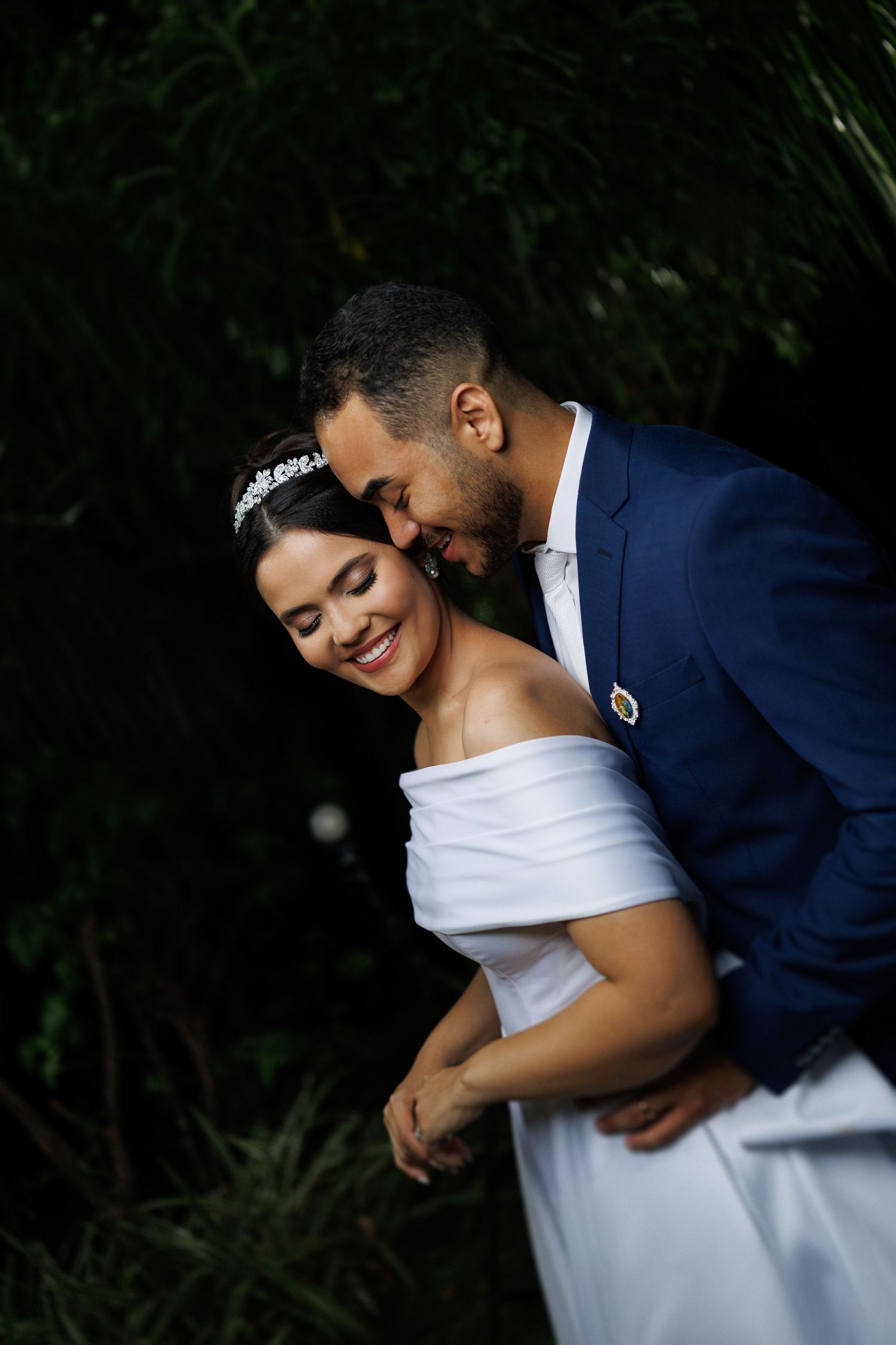 Intimate moment between newlyweds in formal attire against dark foliage.
