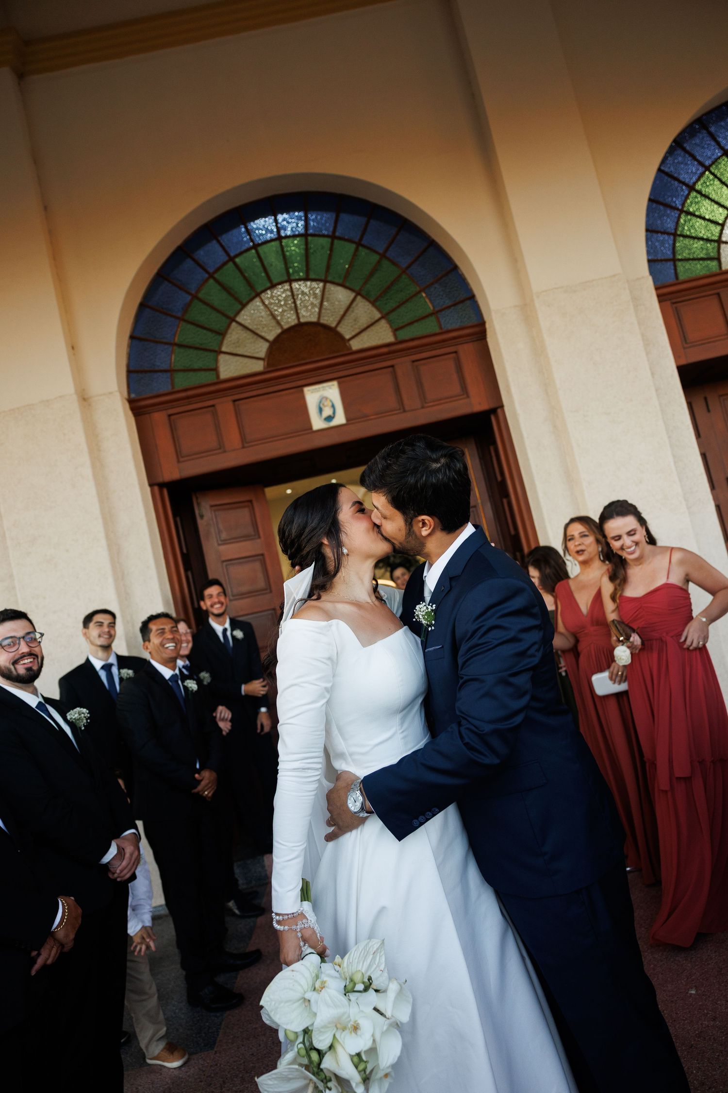 Newlyweds share their first kiss outside a church with wedding party looking on.