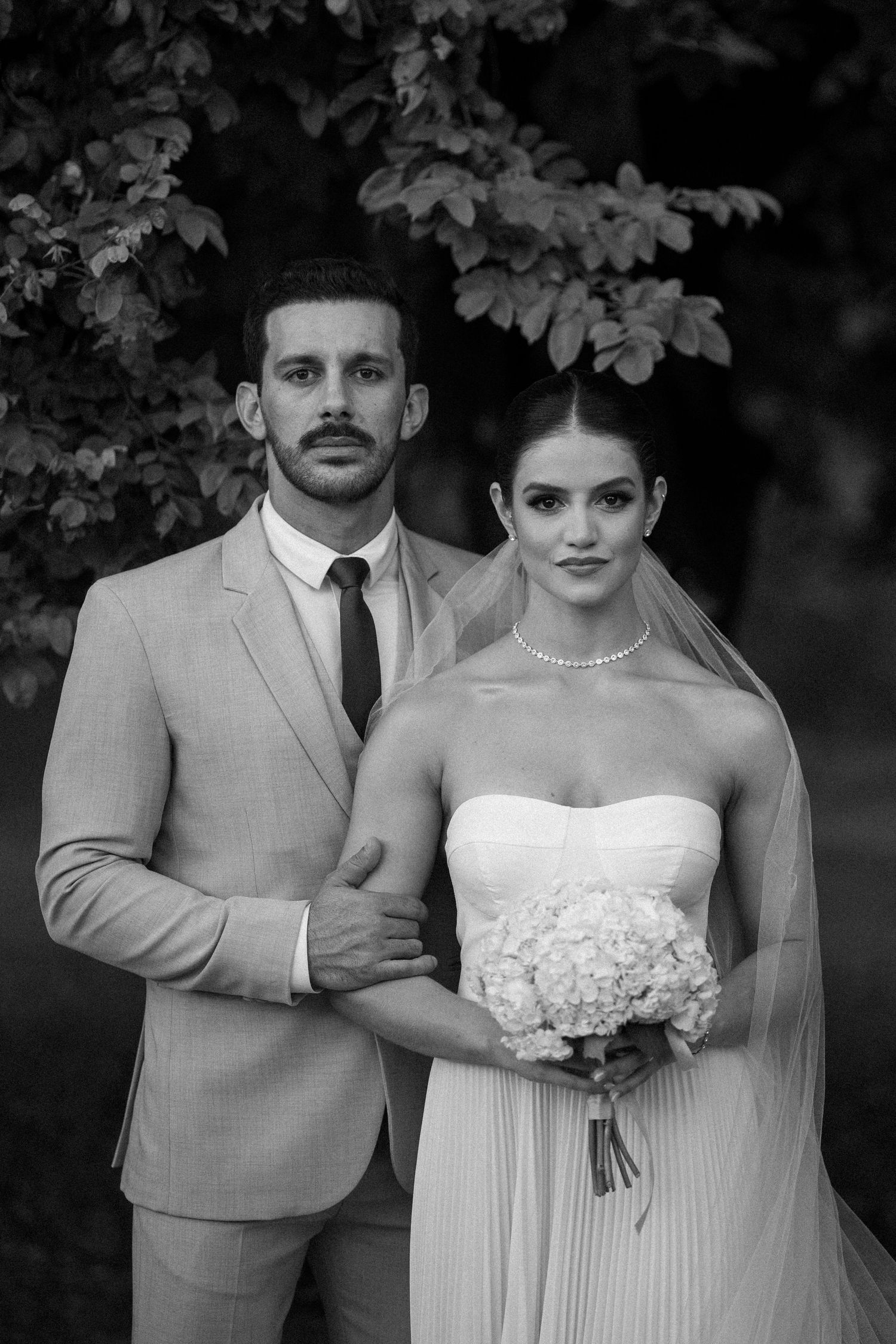 Wedding couple poses in formal attire against a dark foliage backdrop.