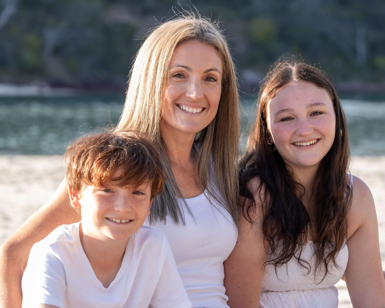 Beach family portrait with mother and her two children sitting on the sand by calm water, warm sunlight and a relaxed coastal lifestyle.