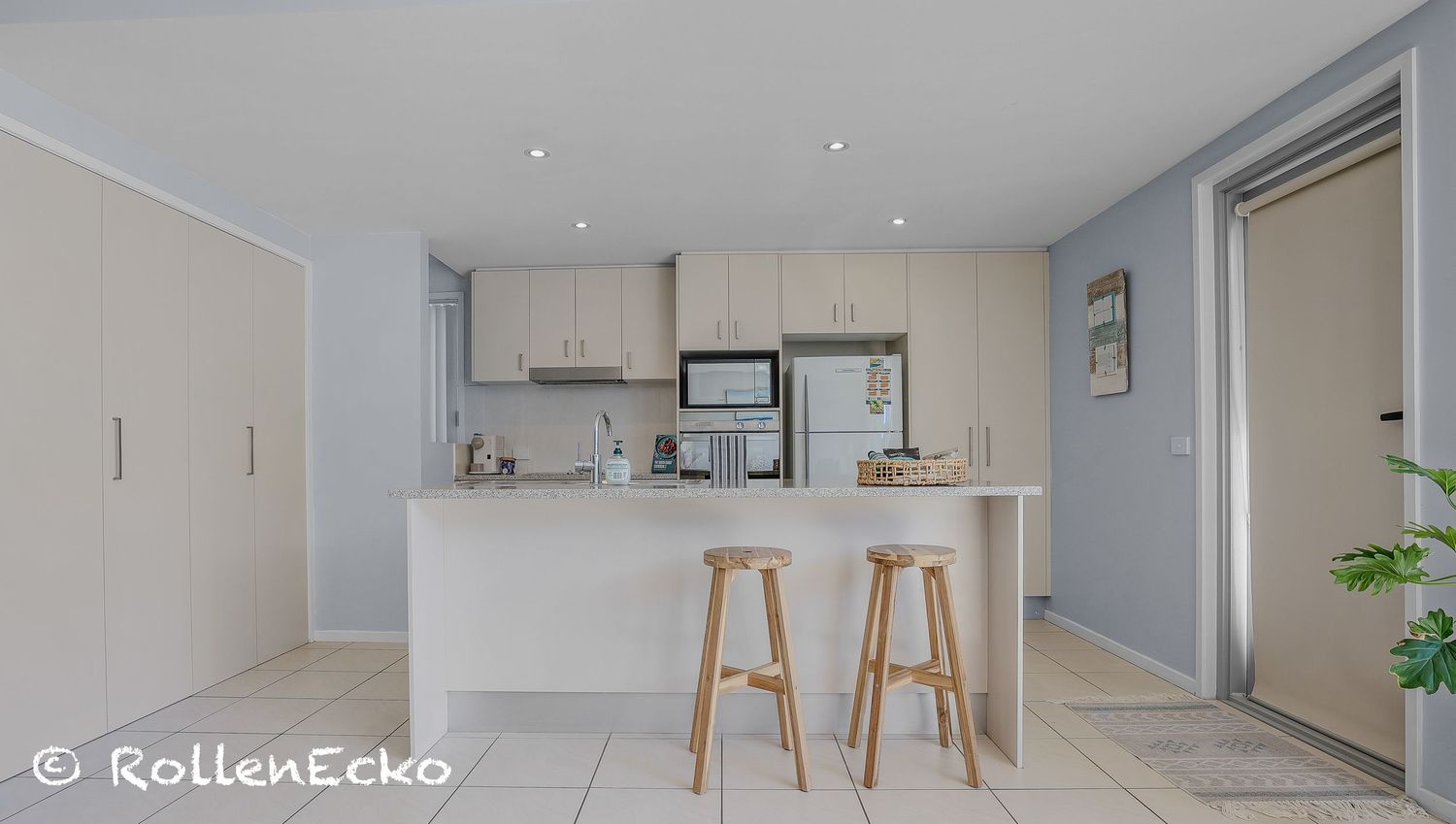 \A bright, modern Airbnb kitchen with neutral cabinets, a stone island bench, two wooden stools, stainless appliances, and soft natural ligh