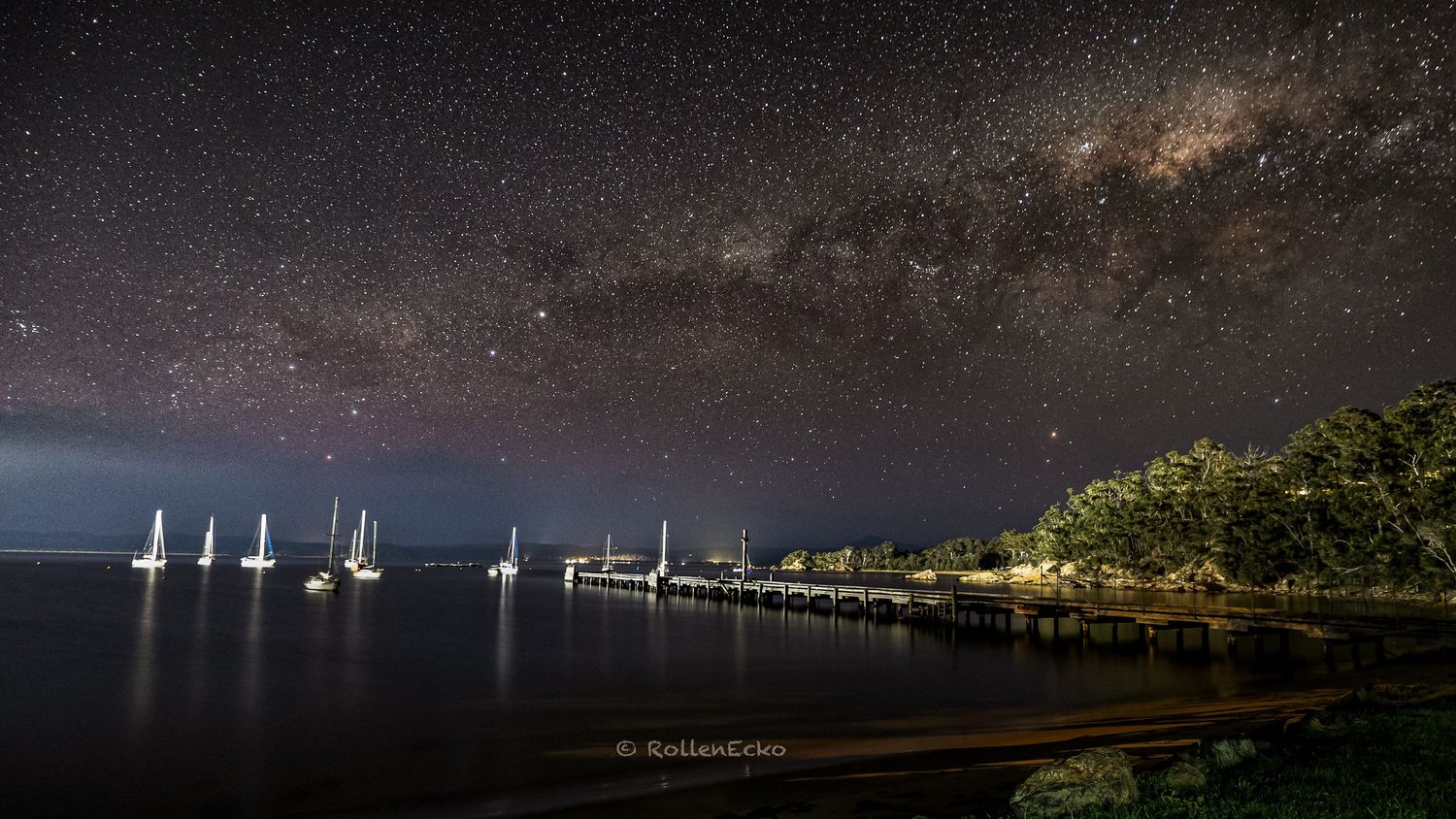 Milky Way above coastal jetty and anchored sailboats, calm water reflections, night sky astrophotography on Australian seaside shoreline