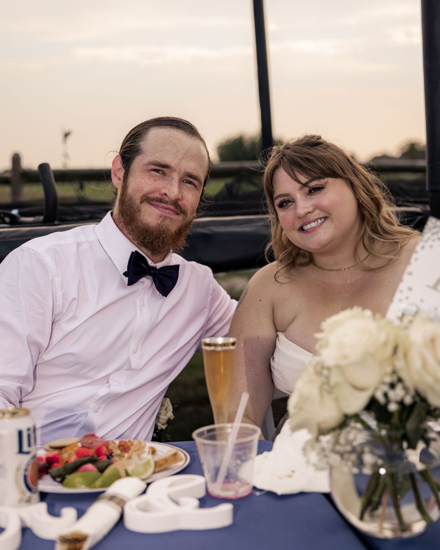A smiling couple in wedding attire sits at a reception table with flowers and food at sunset.