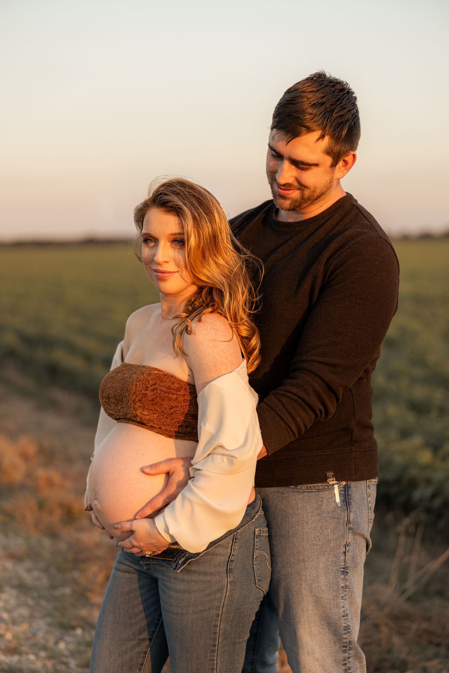 Couple embracing outdoors at golden hour, pregnant woman cradling baby bump in field at sunset.