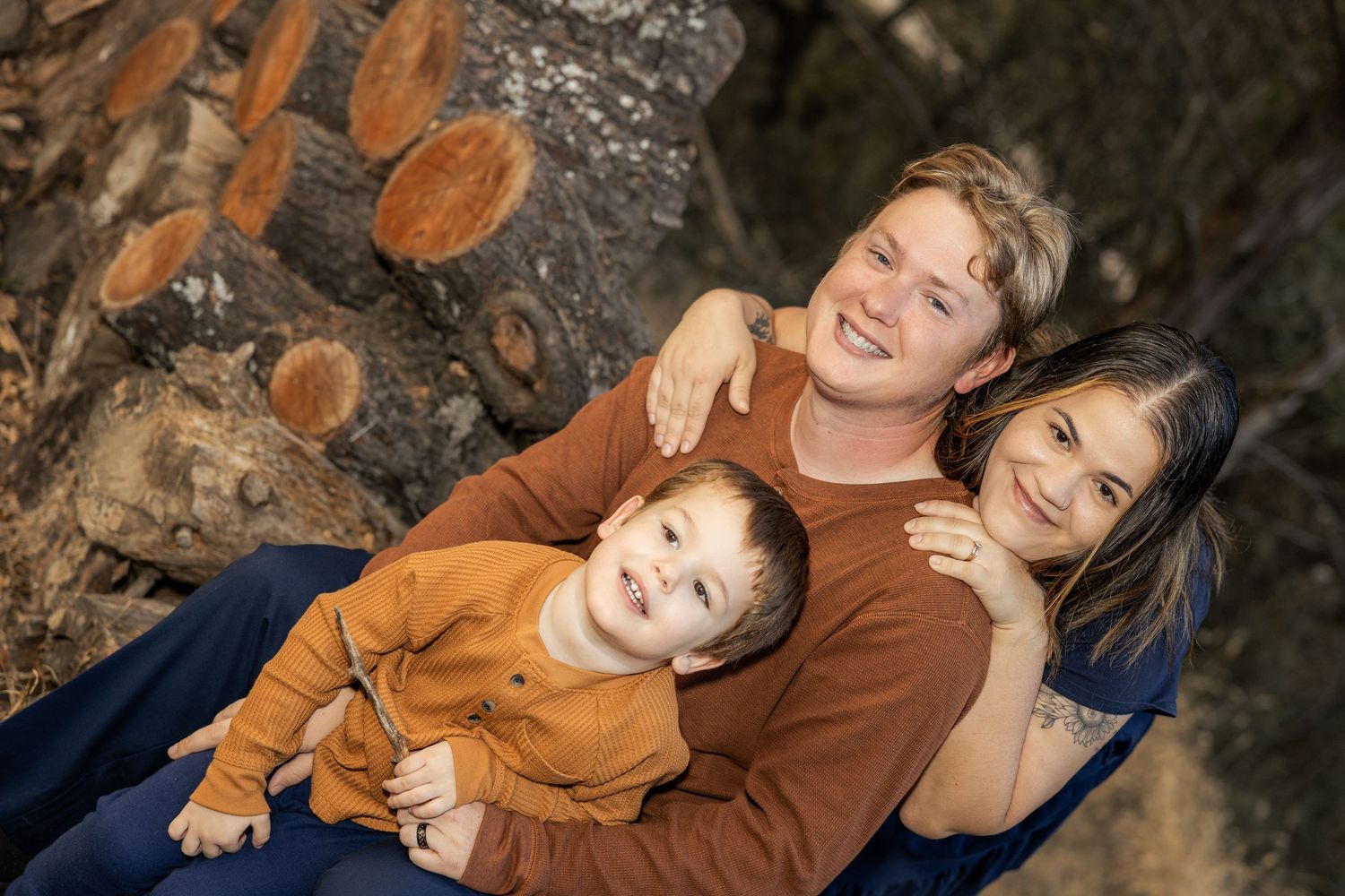 Parents and a toddler boy in matching rust tones smile together outdoors near a rustic log backdrop.