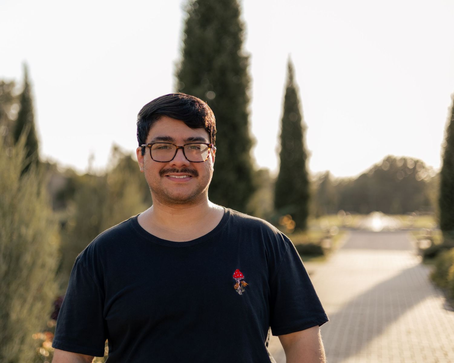 Young man with glasses and mustache smiling outdoors in a sunny park with tall cypress trees in the background.