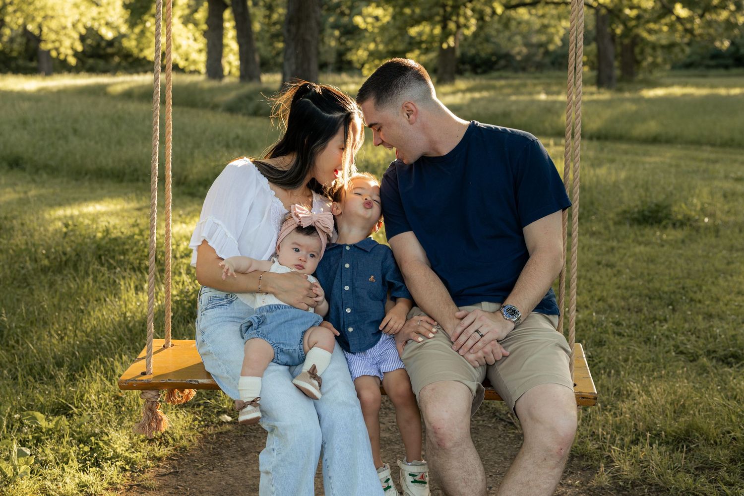 Family of four sitting on a wooden swing in a sunny park, parents smiling at toddler children outdoors.