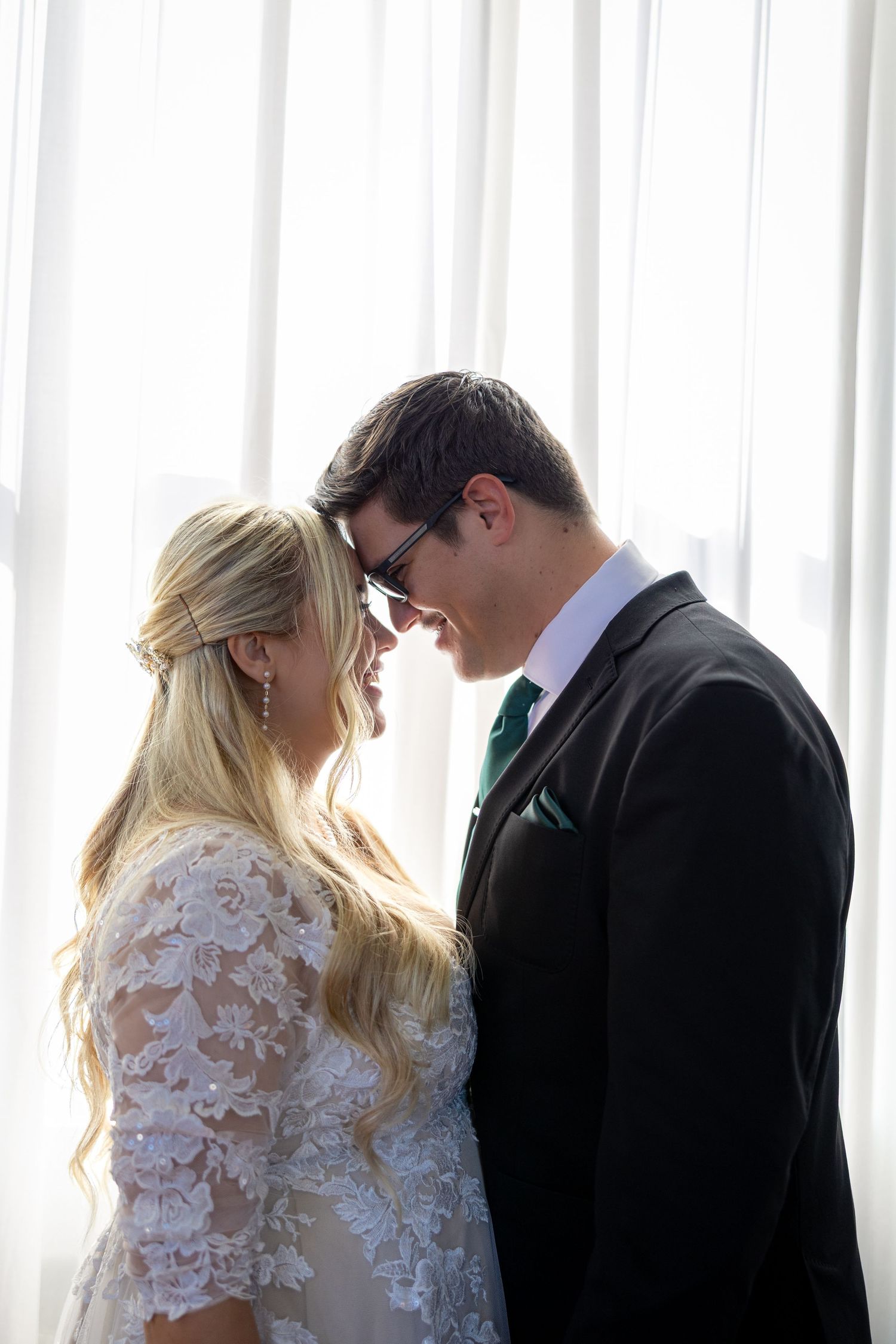 A bride and groom share an intimate forehead touch, smiling softly in front of sheer white curtains on their wedding day.