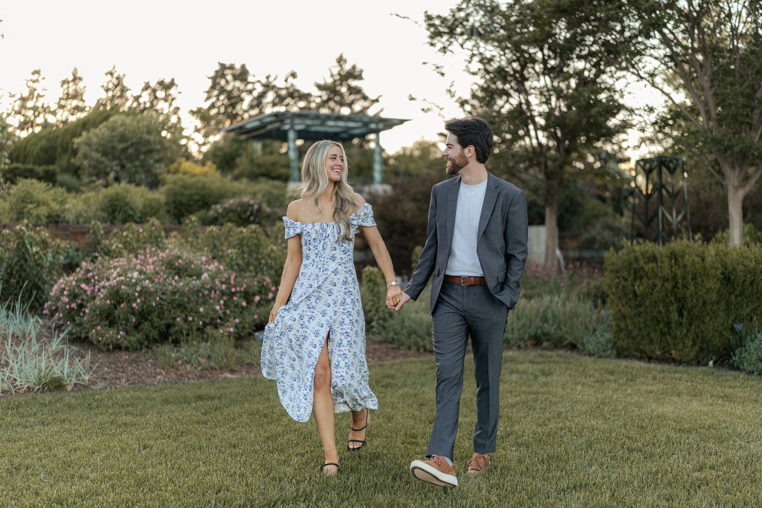 Couple holding hands walking through a lush garden at sunset, woman in floral dress, man in gray suit.