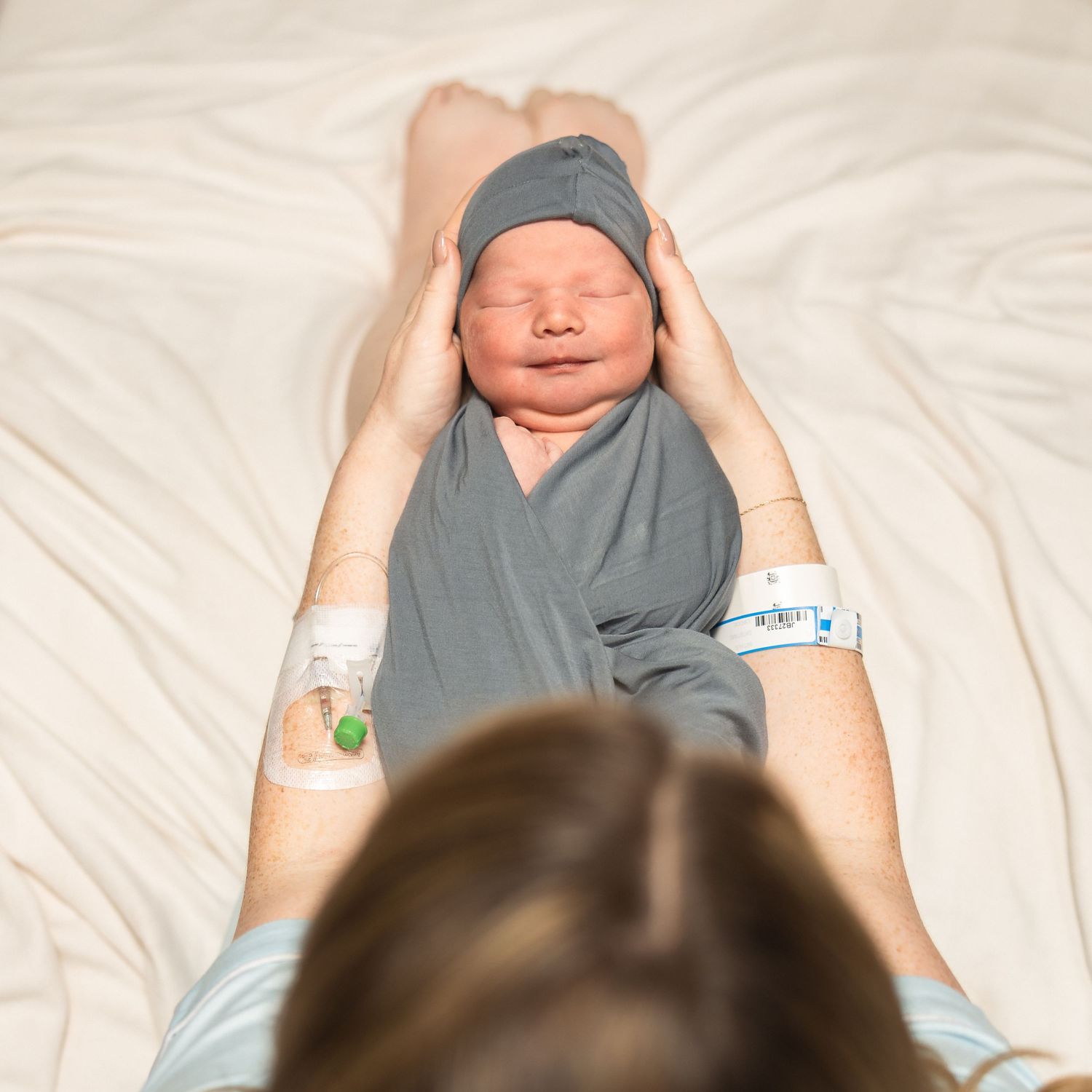 Mother holding swaddled newborn baby in gray hat on white hospital bed after birth.