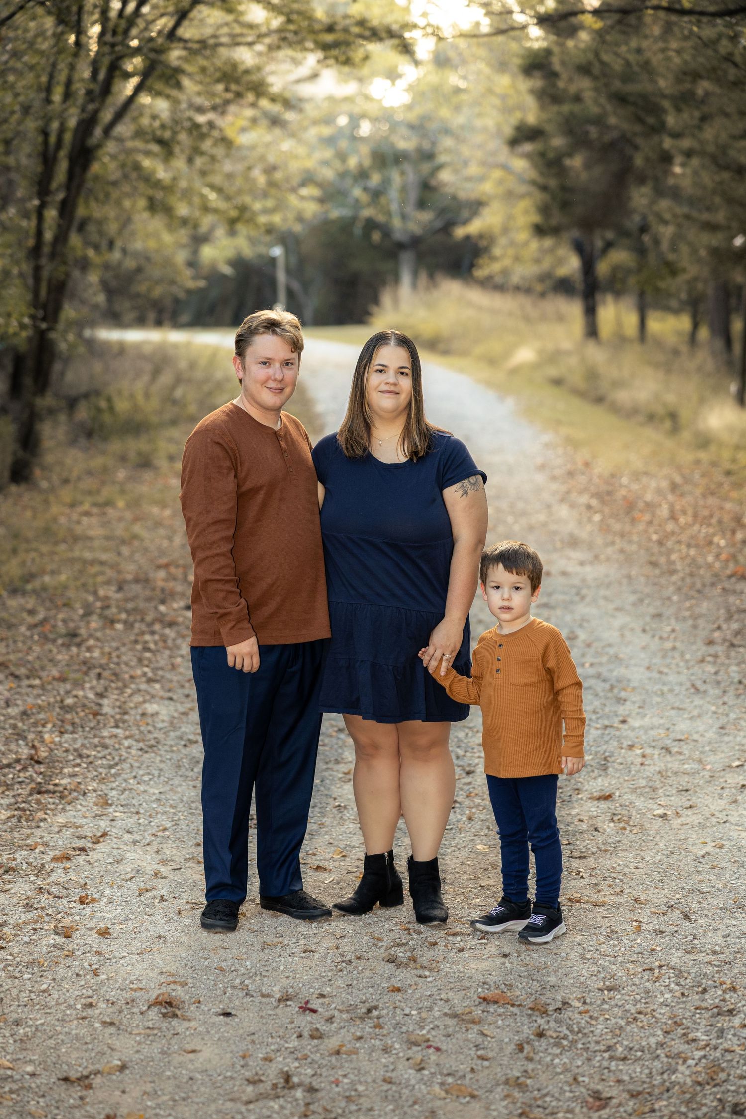 Family of three posing on a scenic gravel path surrounded by autumn trees, wearing coordinated navy and rust outfits.
