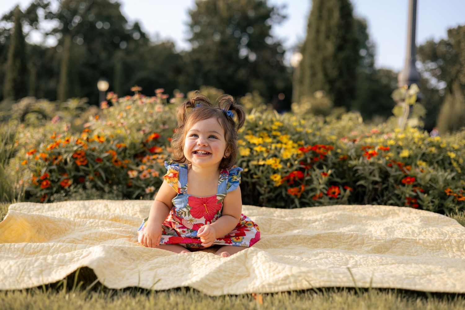 Smiling toddler girl in floral dress sitting on blanket in sunny garden with colorful flowers in background.