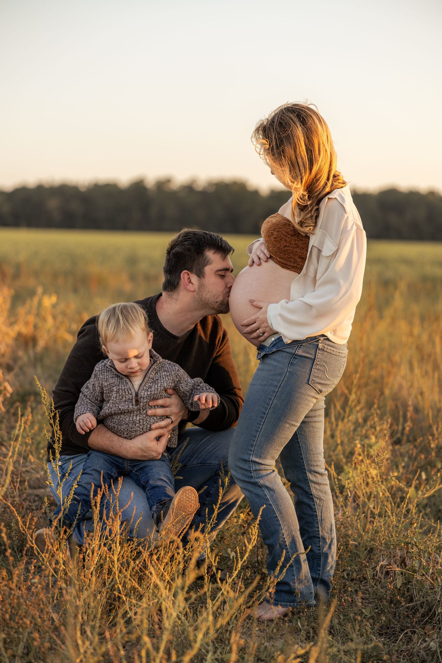 A man kisses his pregnant wife's belly while holding their toddler in a golden field at sunset.