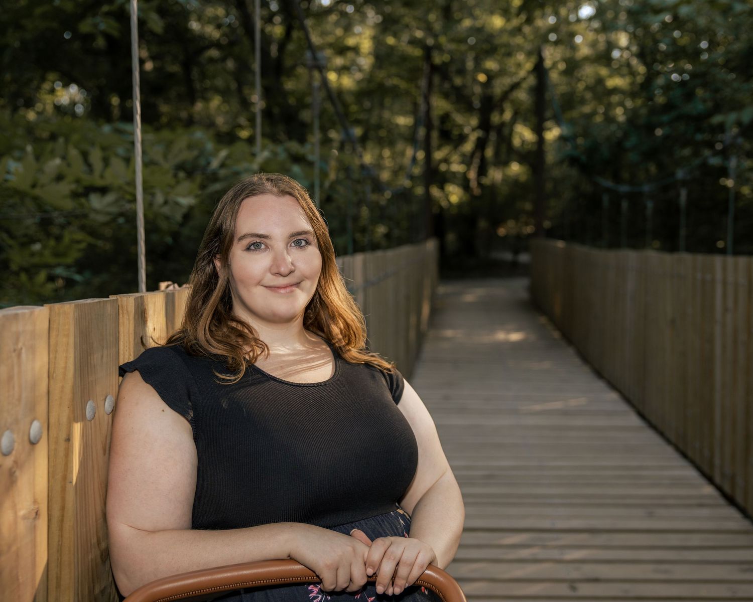 Young woman in black top smiling on wooden bridge walkway surrounded by lush green trees.