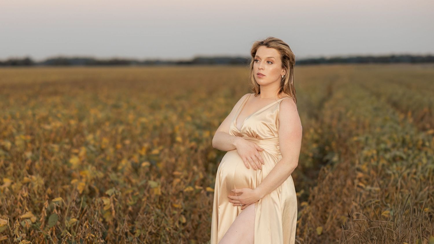 Pregnant woman in a flowing beige dress standing in a green field at dusk, cradling her baby bump.