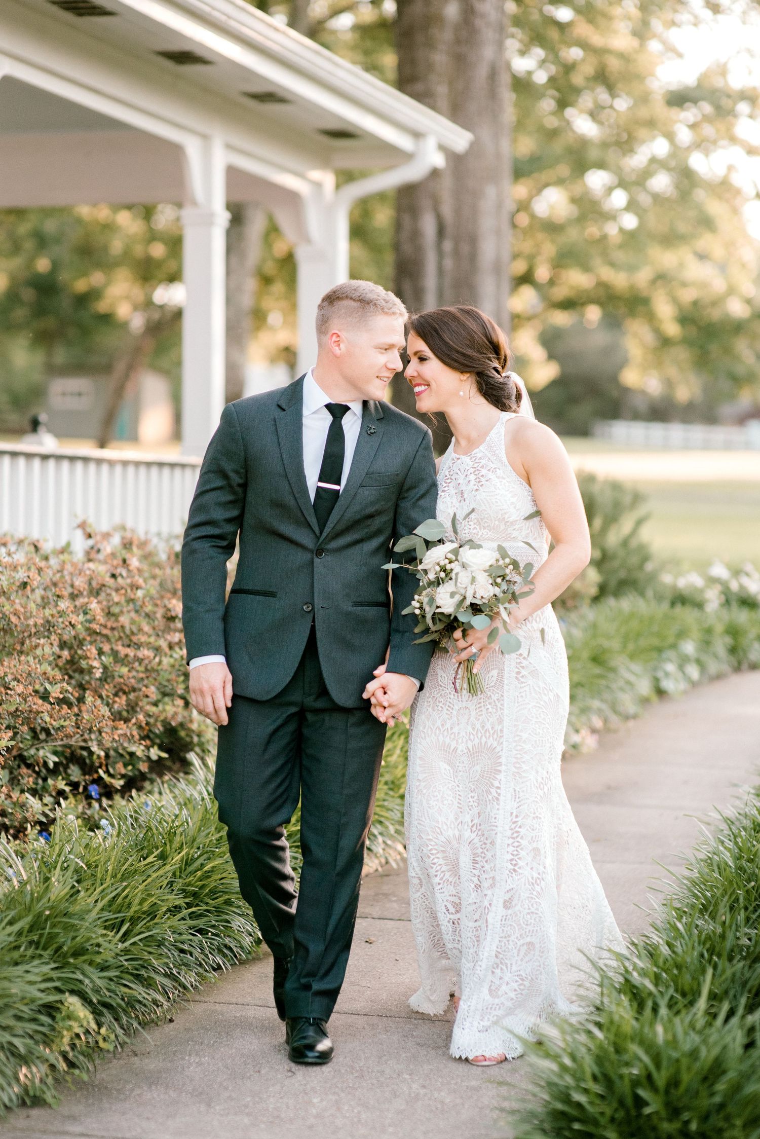 A couple in wedding attire walks together along a garden path lined with green plants on a sunny afternoon.