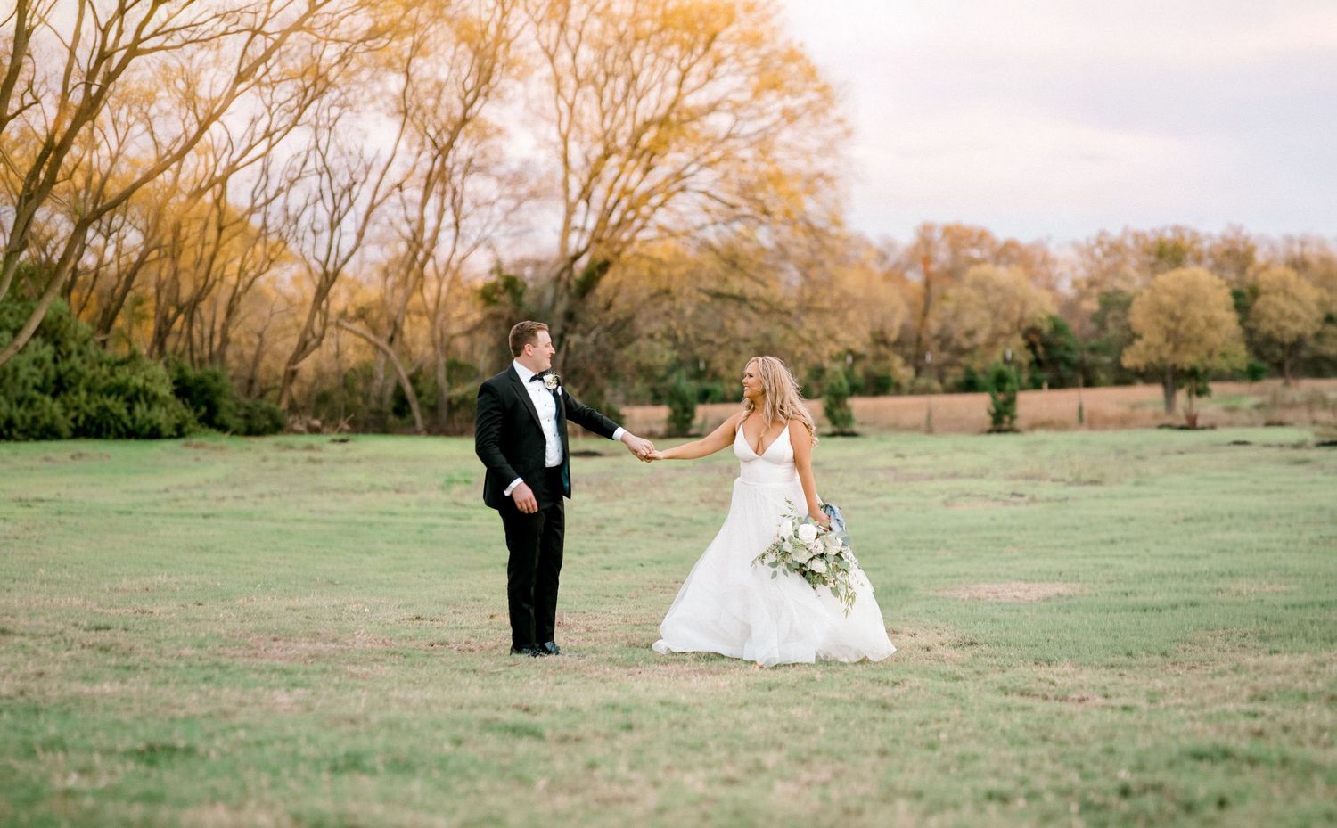 A couple in formal wedding attire stands in a scenic autumn field with golden trees in the background. Wedding Photographer tyler tx