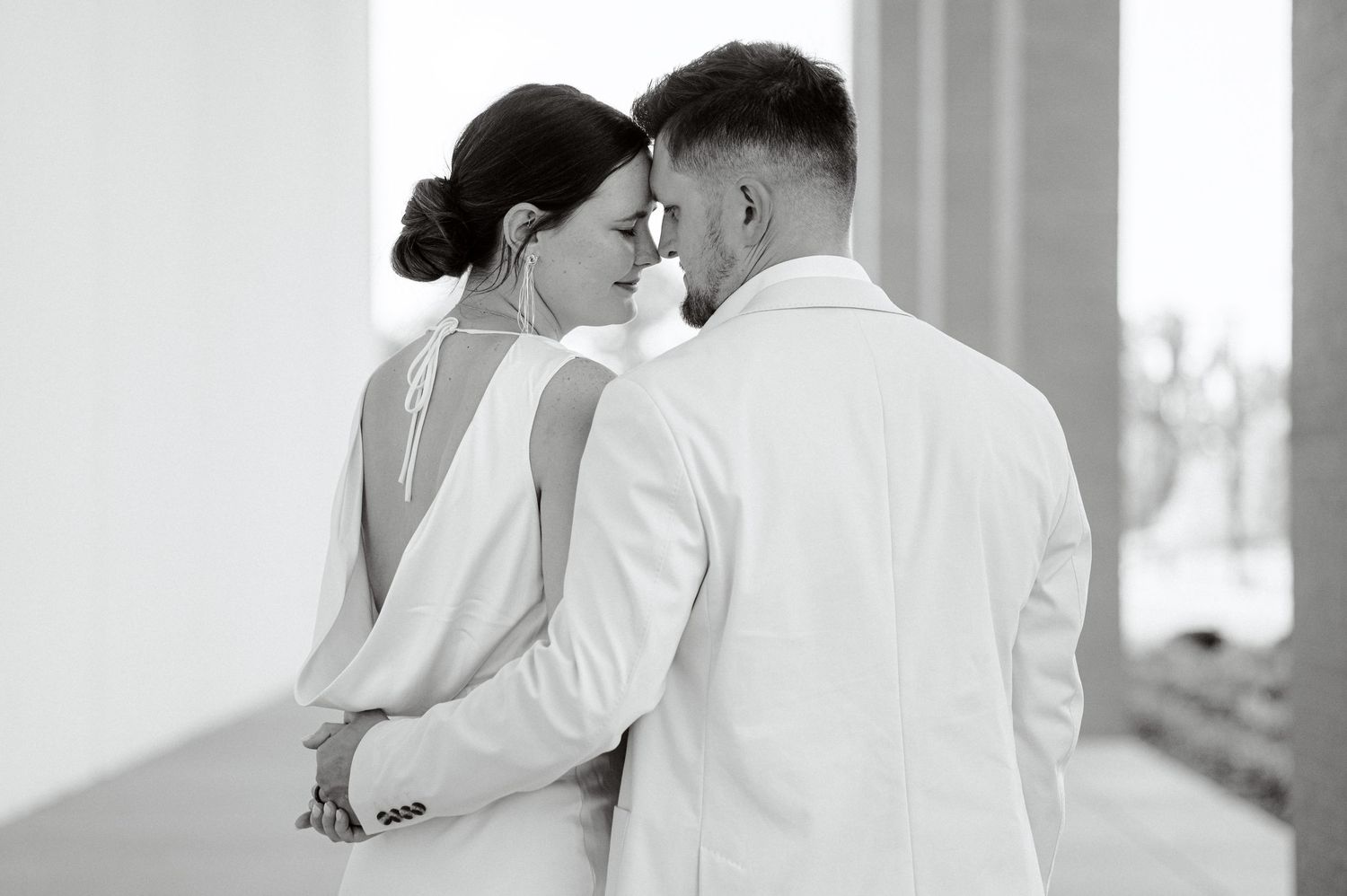 A couple in white wedding attire share an intimate moment while dancing together in a bright room.