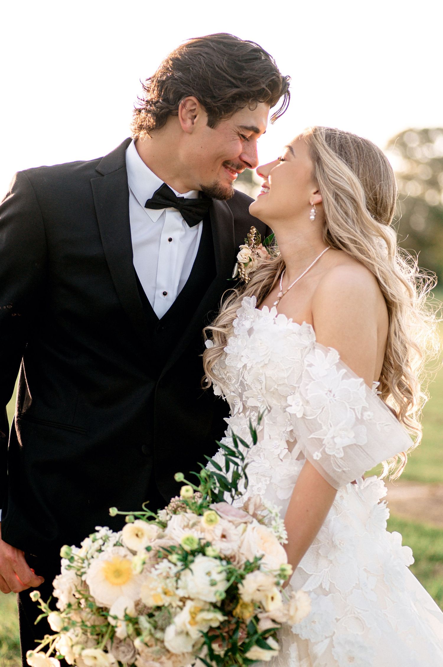 A romantic wedding couple shares intimate moments in formal attire with white floral bouquet during golden hour.