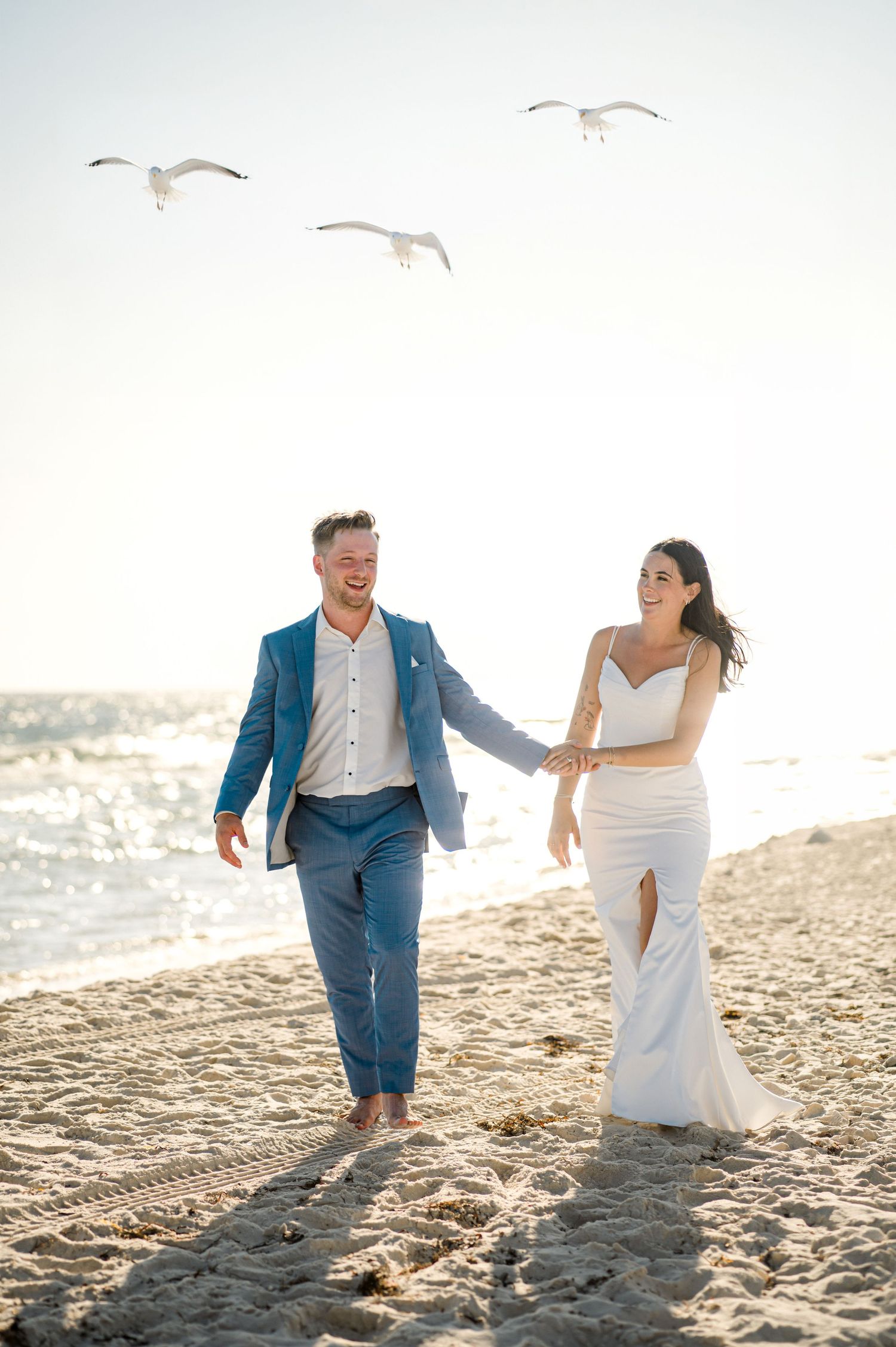 A couple walks hand in hand on a sandy beach at sunset while seagulls fly overhead in the background.